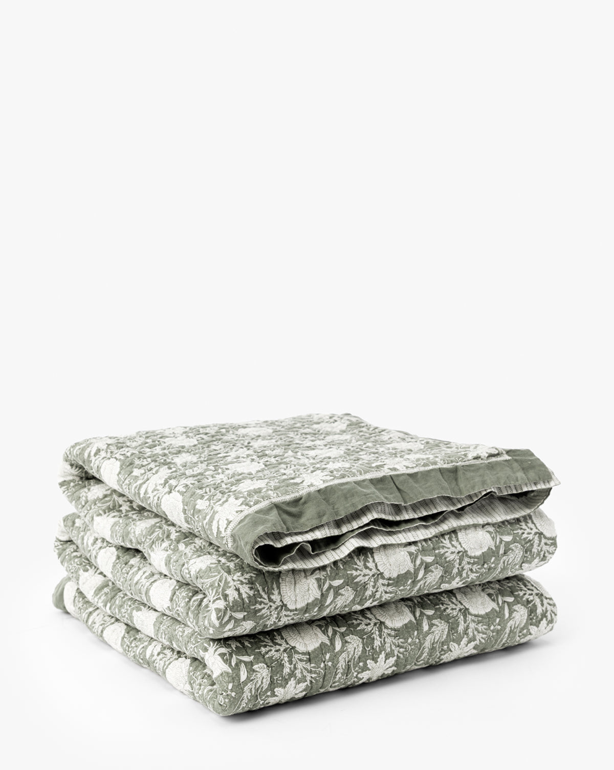 The Audrey Sage Block Print Quilt, featuring a delicate floral pattern and ruffle edging, is neatly folded and stacked on a plain white background by McGee & Co.
