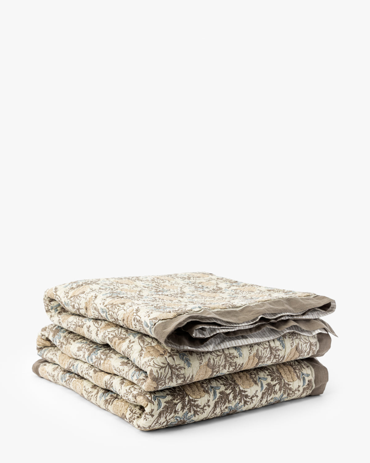 The Audrey Tan Block Print Quilt, featuring beige, brown, and blue floral patterns with delicate ruffle edging, is neatly folded and stacked against a plain white background by McGee & Co.