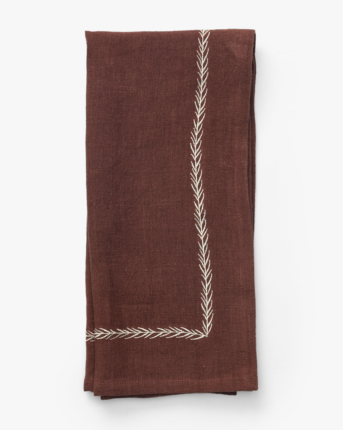 The Broadleaf Tea Towel is a folded brown linen piece featuring an embroidered leaf border at the corner, perfect for adding elegance to your kitchen, shown on a plain white background by McGee & Co.