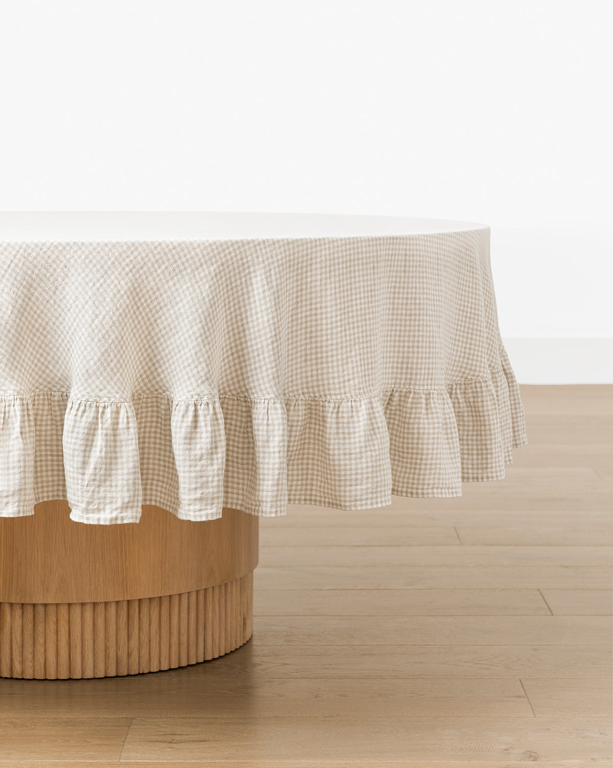 A round wooden table is covered with the Catania Linen Ruffle Tablecloth in beige and white gingham, set on a light wood floor against a plain white wall, by McGee & Co.