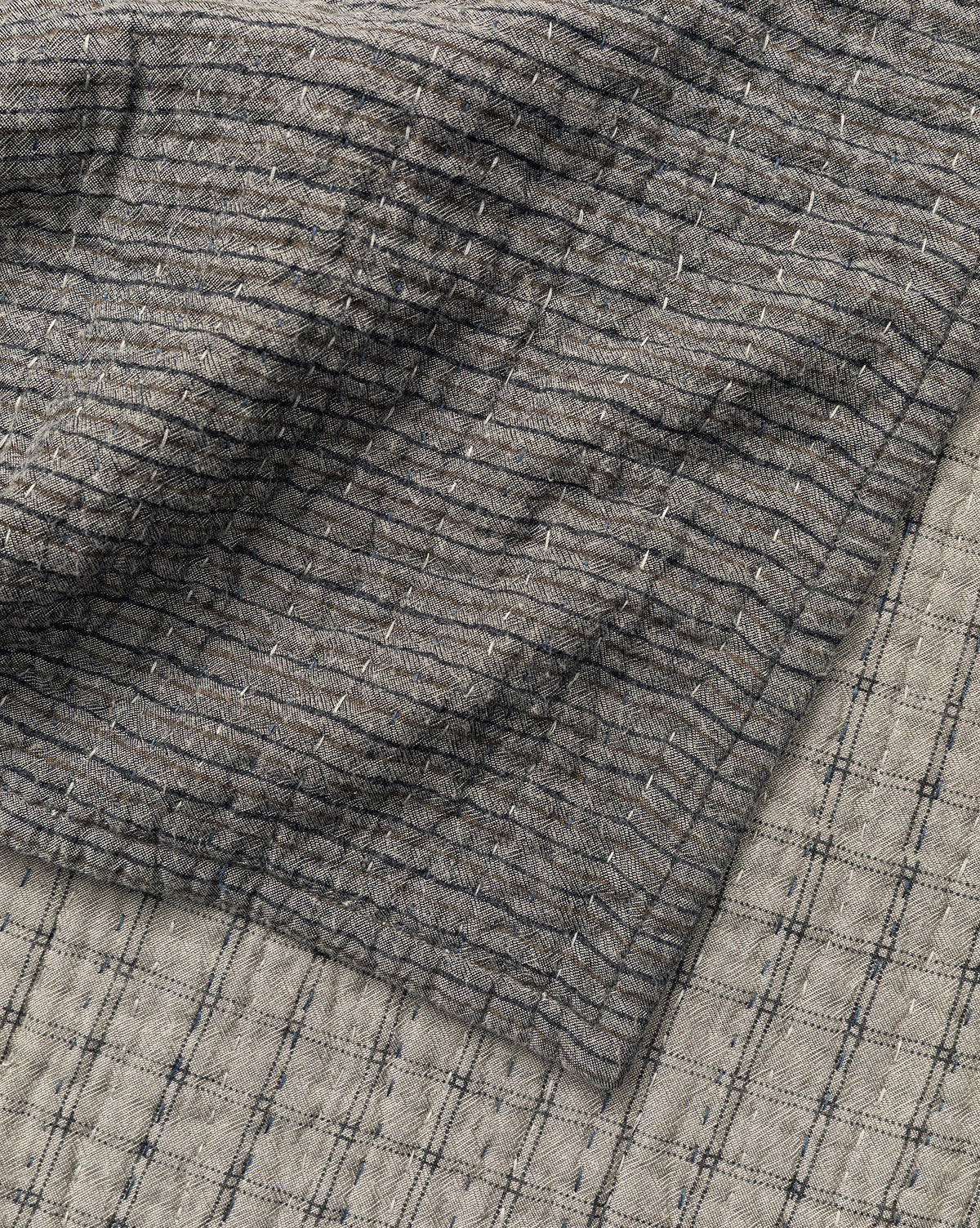 A close-up of two overlapping Cecily Kids Quilts by Renuka Inc. shows textured, slightly wrinkled fabrics: the top features a darker grid pattern with fine lines, while the bottom displays a lighter shade with bold grid lines—McGee & Co.