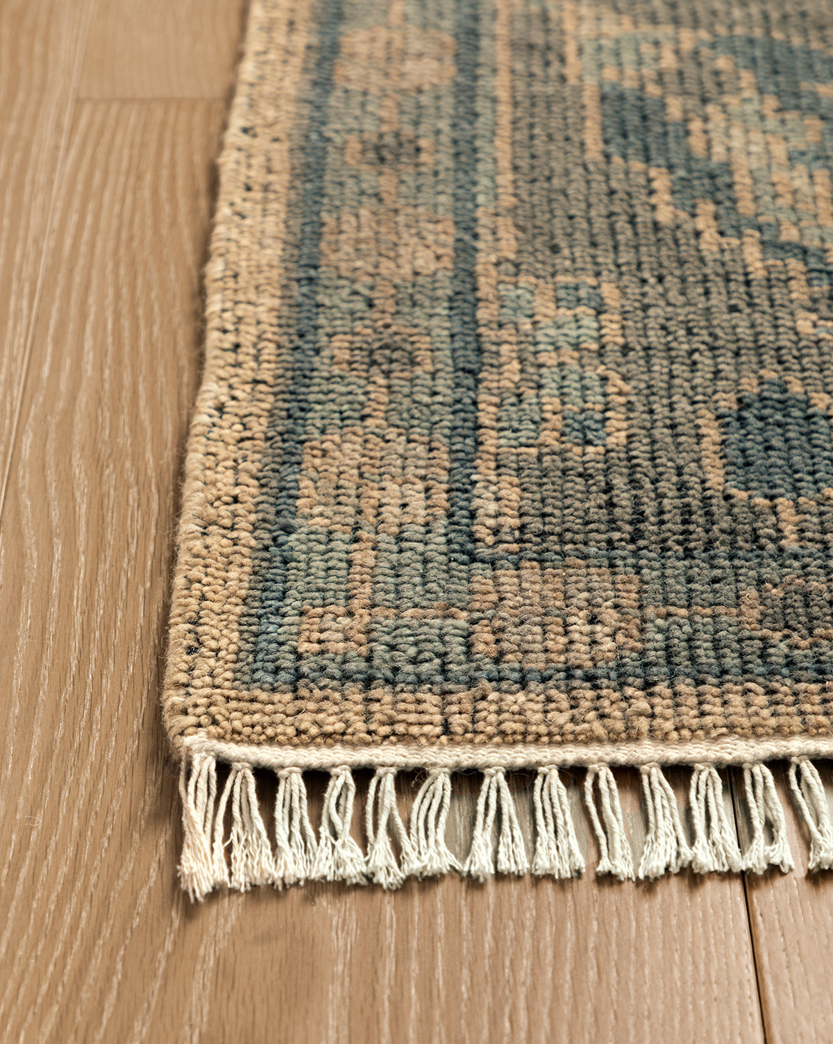 A close-up of the Chevalier Hand-Knotted Wool Rug displays its blue and beige pattern, fringed edges, and timeless elegance as it rests on light brown wooden flooring, by McGee & Co.