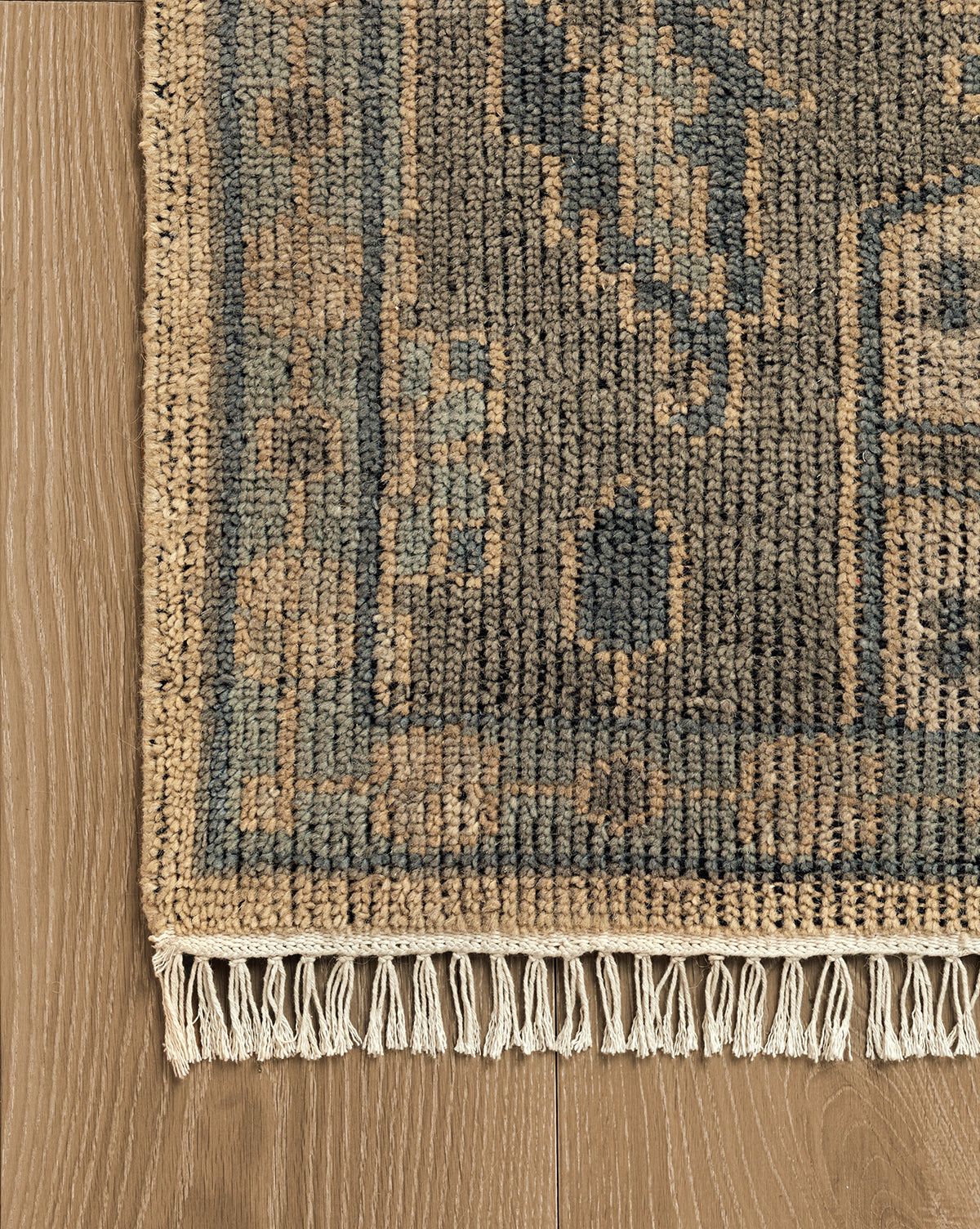 A close-up of the Chevalier Hand-Knotted Wool Rug, featuring a blue background with tan geometric patterns and beige fringe, resting on a light brown wooden floor, showcases its timeless elegance, by McGee & Co.