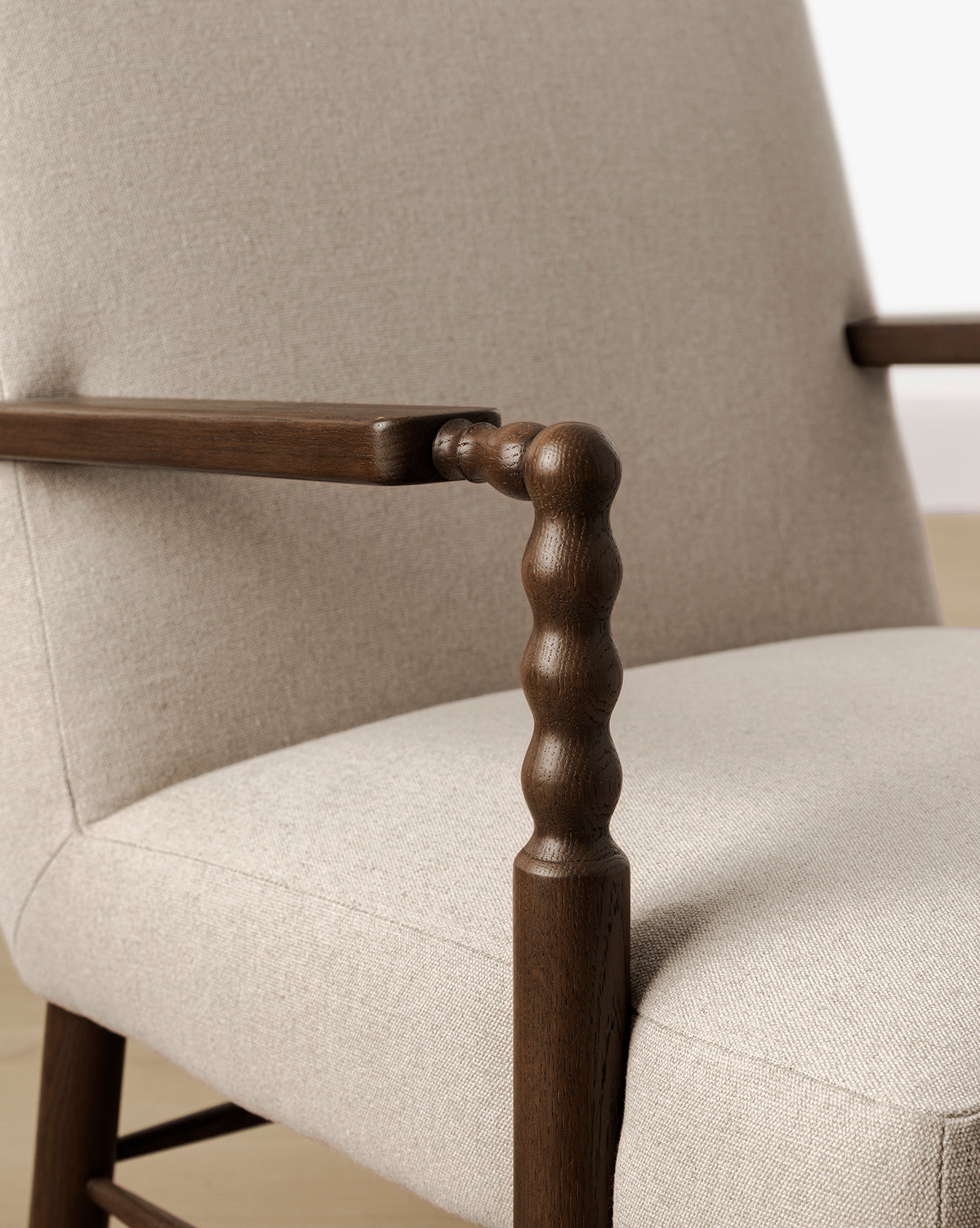 Close-up of the Earnshaw Lounge Chair showcasing neutral-toned, textured upholstery with dark wood armrests and legs in an English oak finish, by McGee & Co. Foreground displays a rounded spindle armrest design set against light-colored flooring and walls.