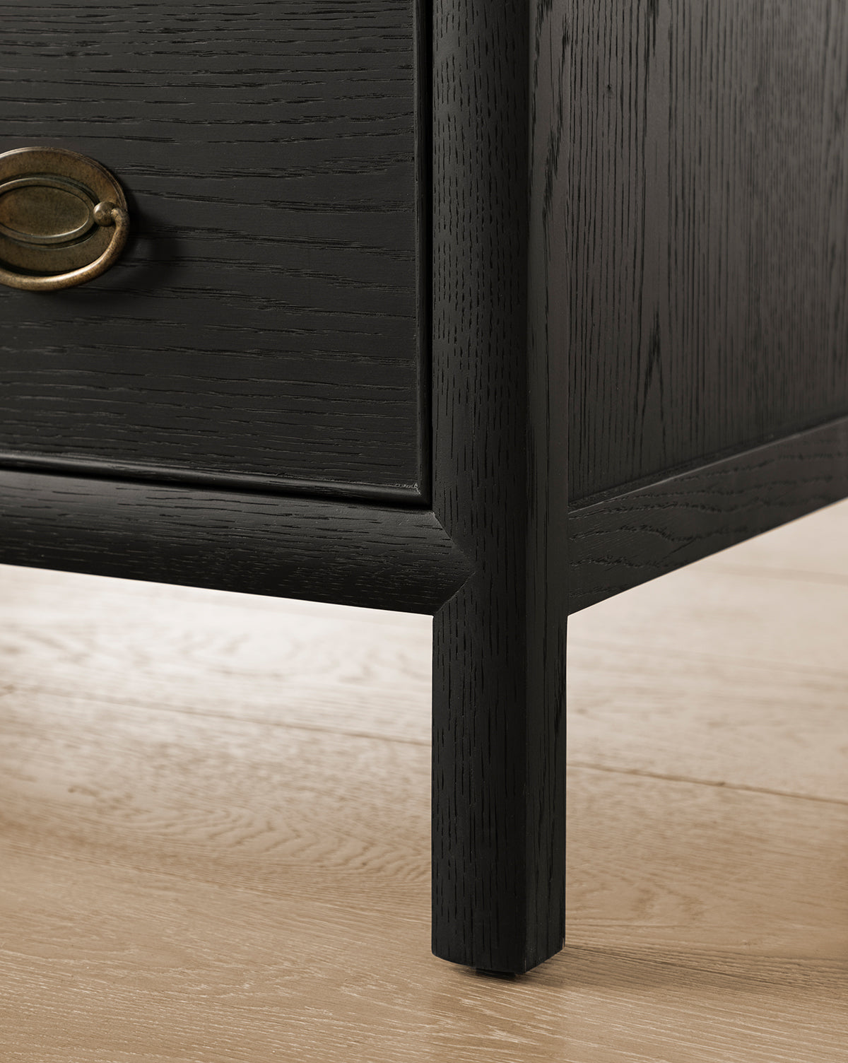 Close-up of the Edward Dresser’s lower corner, showcasing its dark Black Ash finish and brass handle on a light wood floor. The image highlights the textured grain—an elegant bedroom storage solution, by McGee & Co.