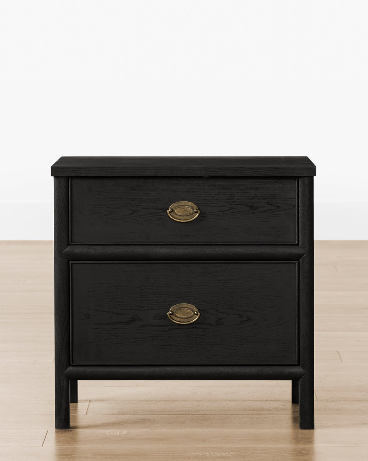 The Edward Nightstand is a black wooden bedside table with two drawers and brass oval handles, shown on a light wood floor against a plain white wall by McGee & Co.