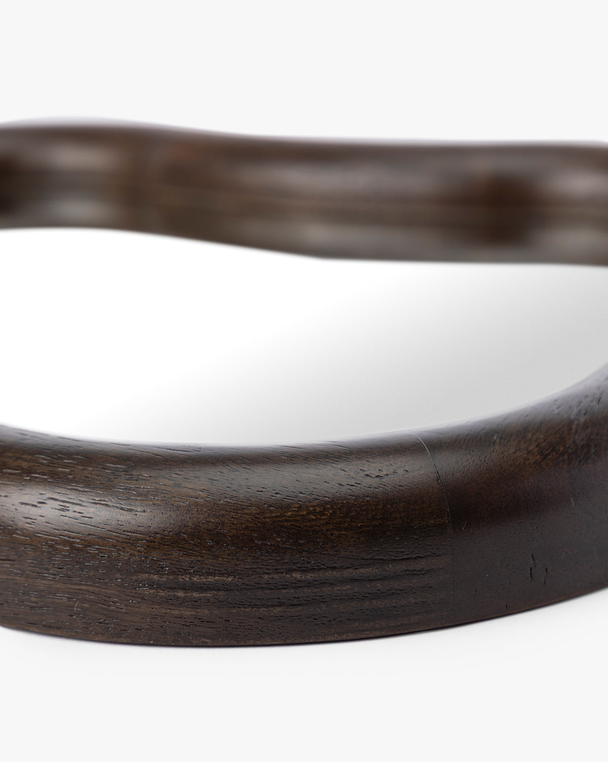 Close-up of the Freya Shelf Mirror’s dark, smooth, curved wooden frame with visible wood grain against a white background—an elegant entryway accent with part of the reflective surface shown by McGee & Co.