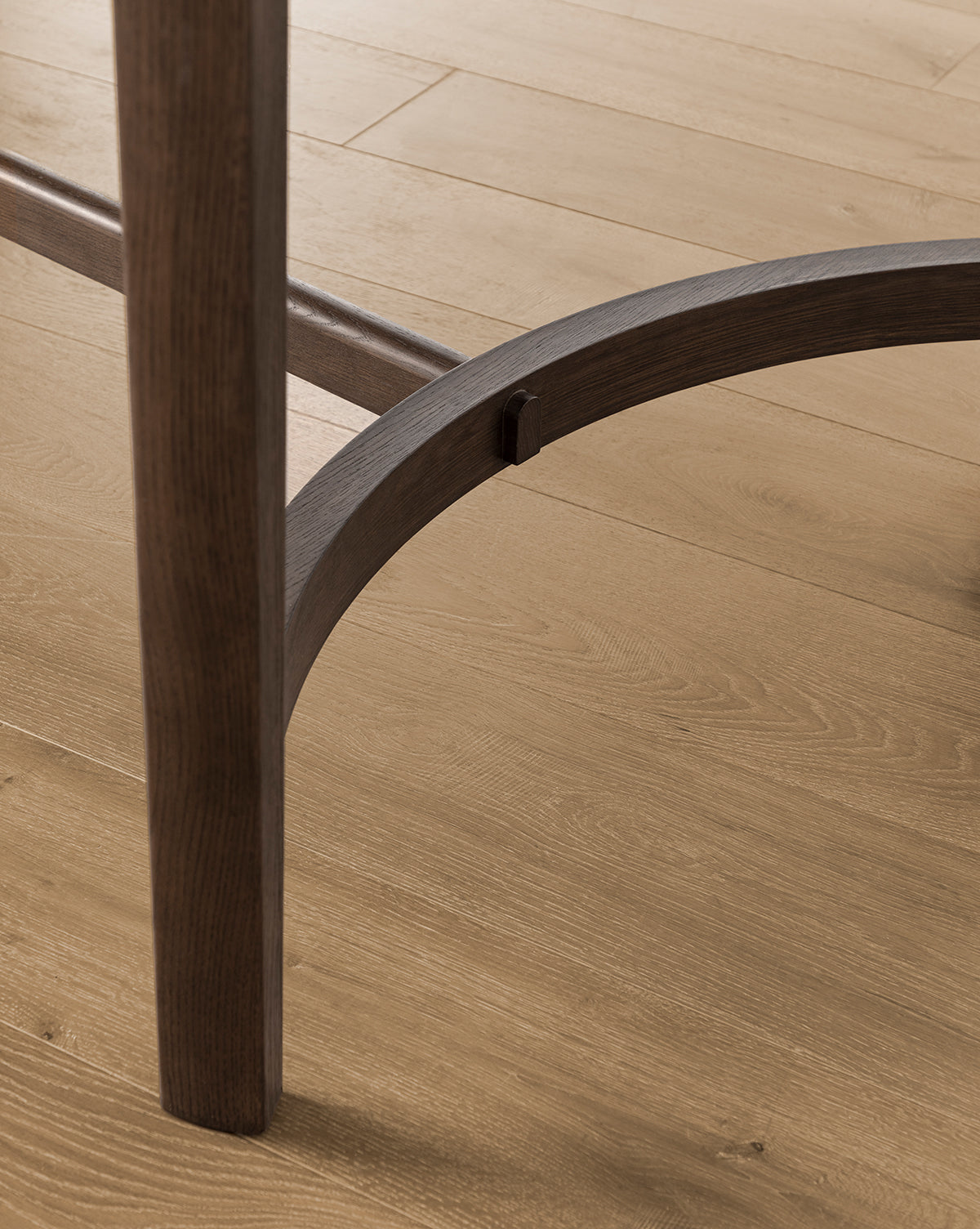 Close-up of the Gardiner Dining Table legs and curved support beam on a light wood floor, highlighting the Walnut Oak grain and texture featured in both the table and flooring, by McGee & Co.