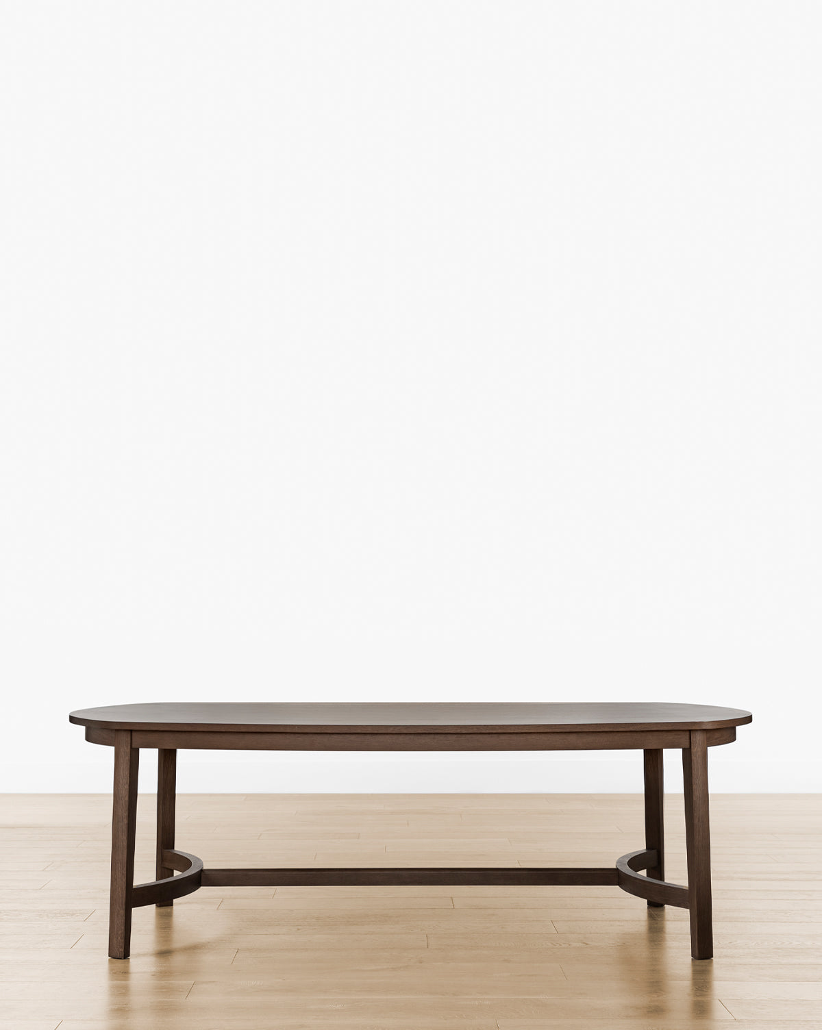 The Gardiner Dining Table, featuring a dark walnut oak oval design with four legs and a low crossbar base, stands elegantly on a light wood floor against a plain white background, by McGee & Co.