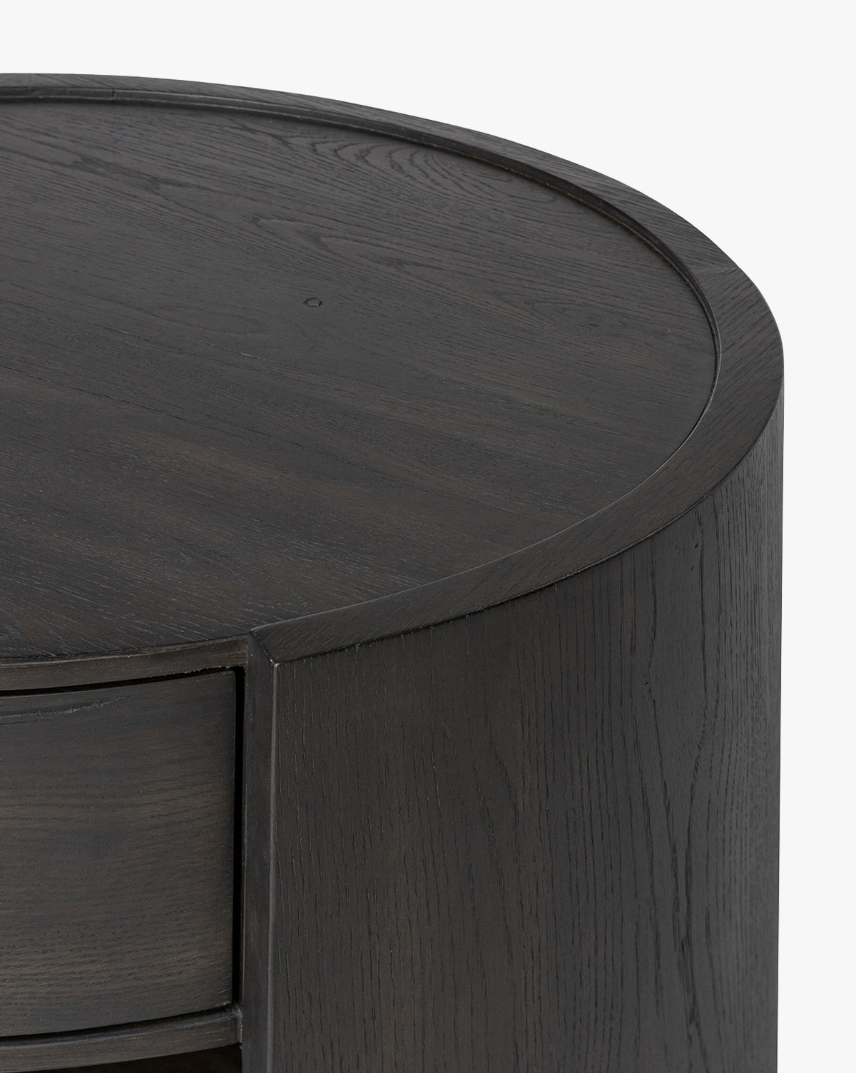 Close-up of the top and side of the Glenndon Oak Nightstand, a round piece with a black finish, smooth surface, and visible wood grain. A drawer is partially visible on the left side, by McGee & Co.