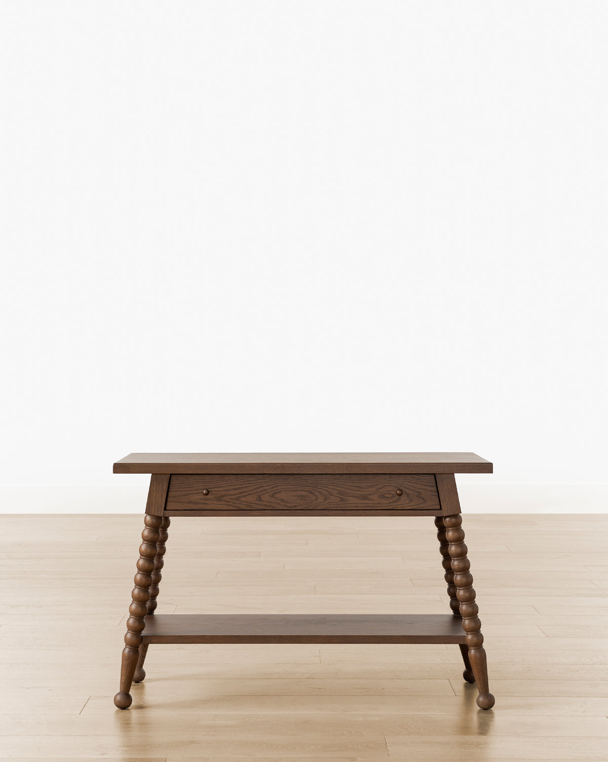The Heathcliff Console, a wooden side table with a walnut oak finish, single drawer, lower shelf, and spiral-turned legs, stands on a light wood floor against a plain white background, by McGee & Co.