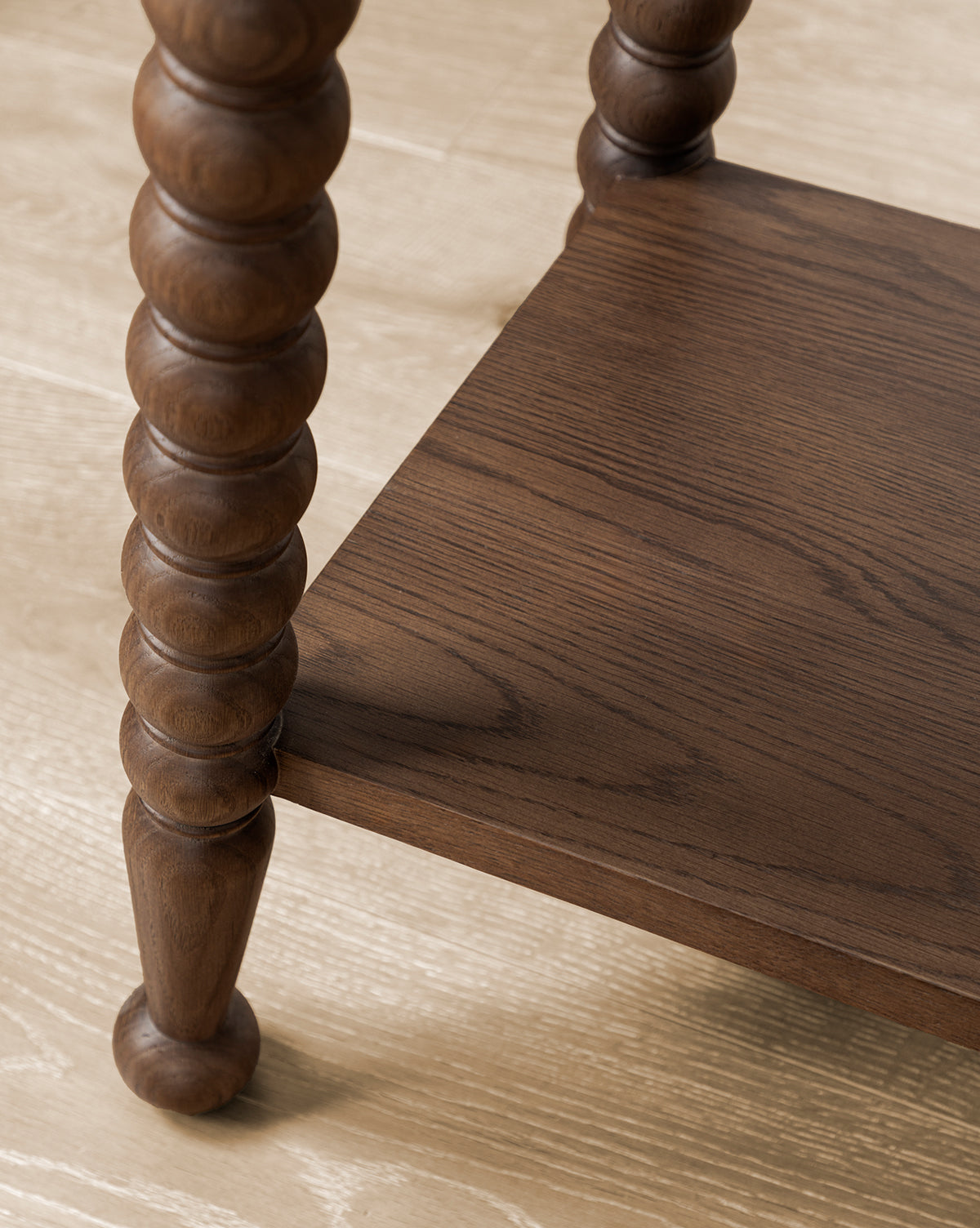 A close-up view of the lower shelf and leg of the Heathcliff Console, featuring round carved bobbin detailing and a walnut oak finish, set on a light wood floor, by McGee & Co.