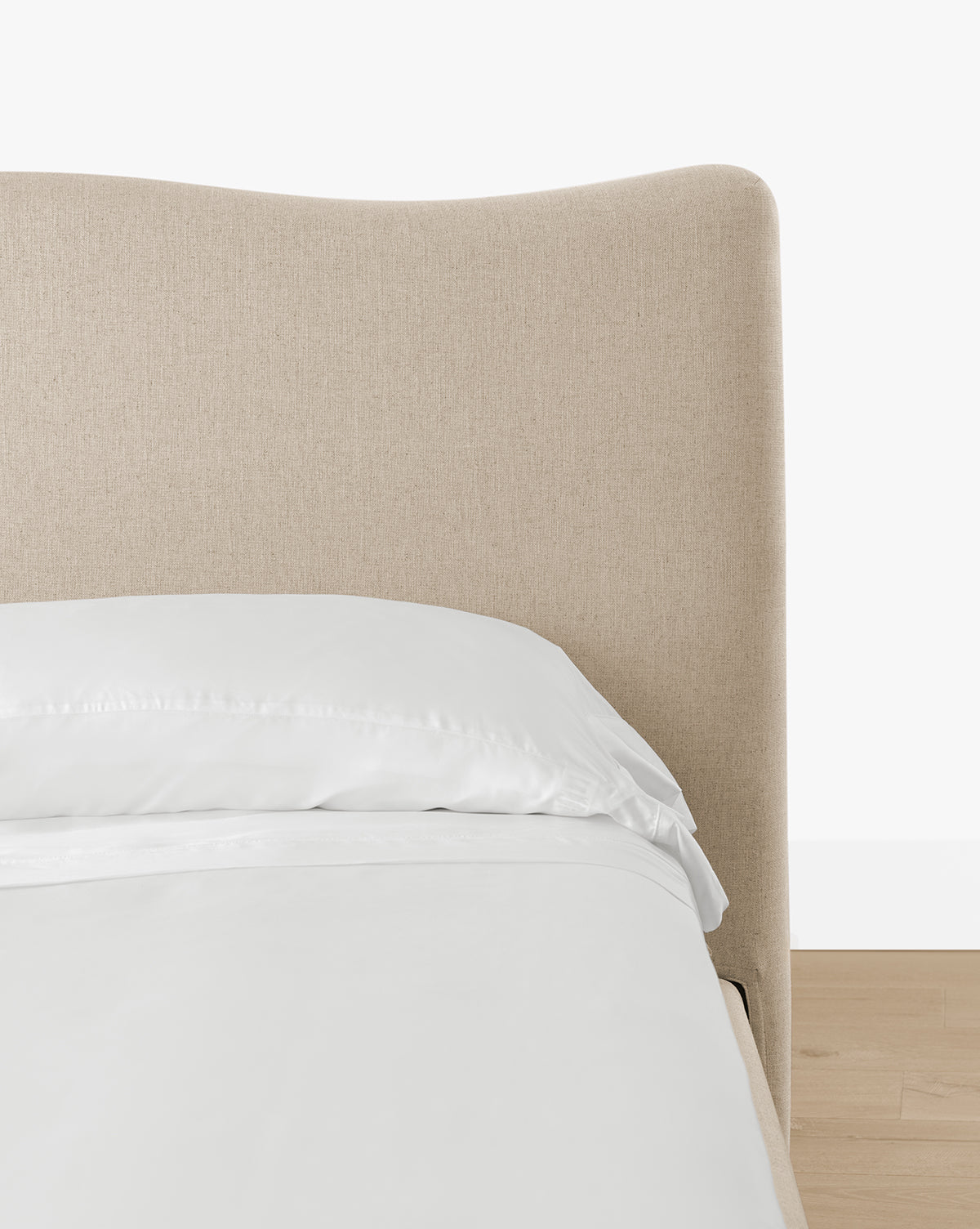 A close-up of the Henrietta Bed (Ready to Ship) featuring a light beige curved headboard, white pillow, and sheets on a light wood floor against a plain white background—perfect for modern bedroom styling, by McGee & Co.
