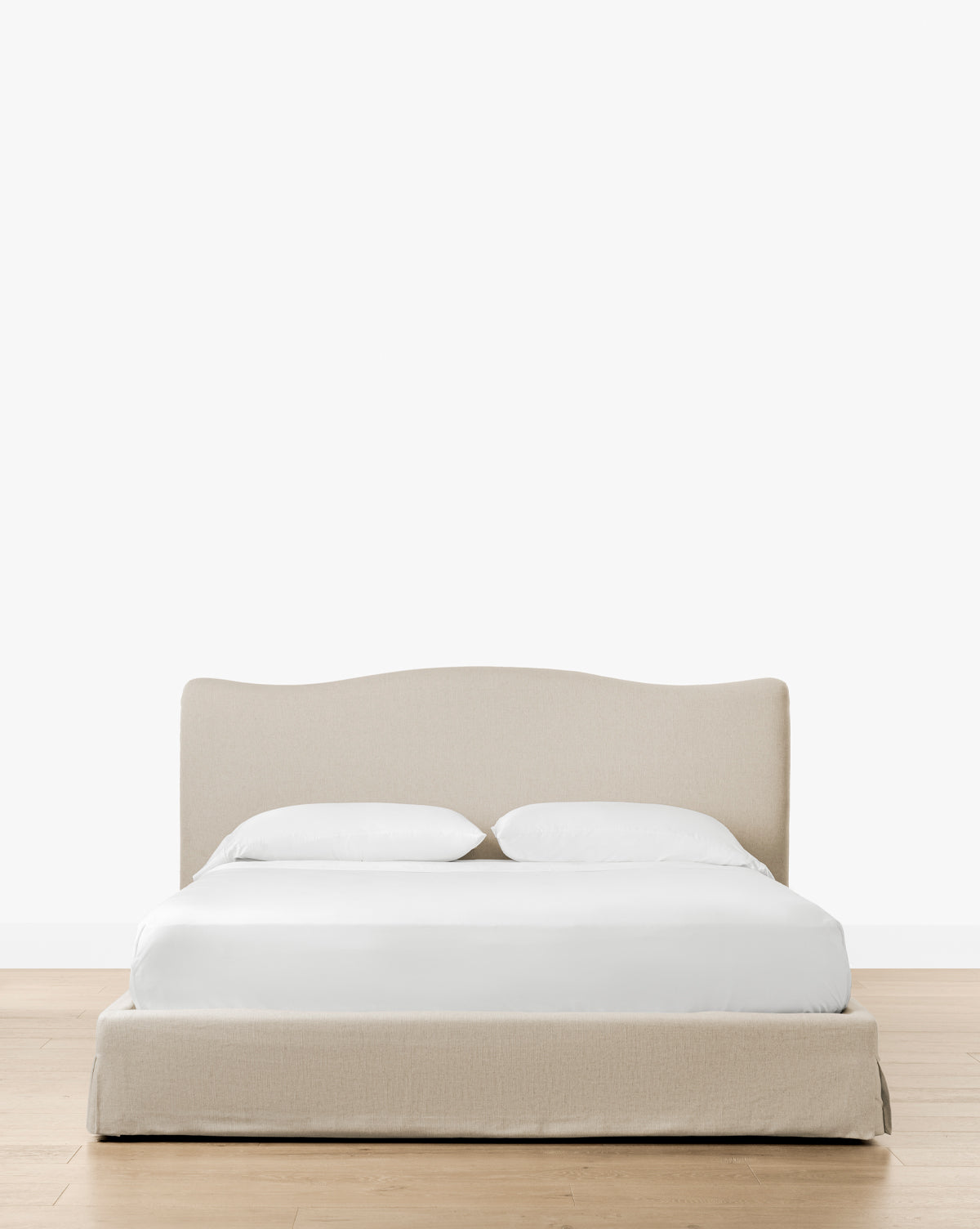 The Henrietta Bed (Ready to Ship), featuring beige upholstery, a curved headboard, white bedding, and two pillows, sits on a light wood floor with a plain white backdrop—ideal for refined bedroom styling by McGee & Co.