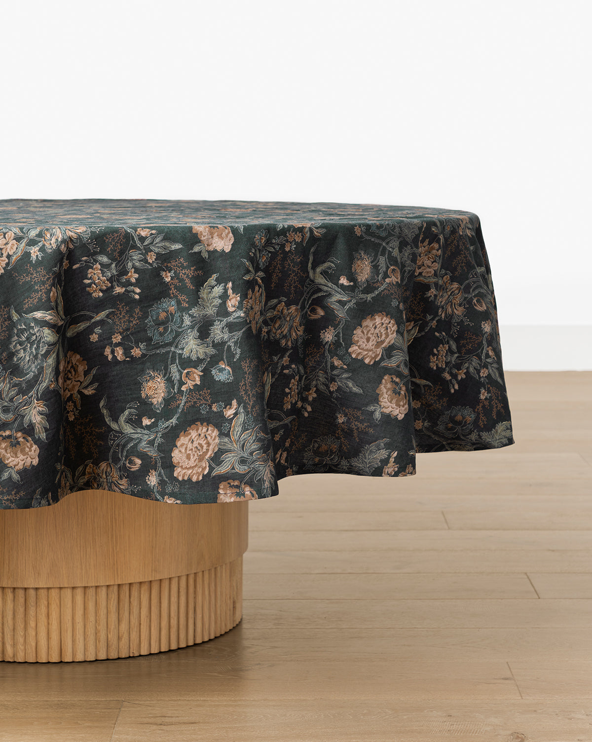 A round table with a light wood base is draped in the Hestercombe Tablecloth, showcasing vintage-inspired muted pink and green florals. The table sits on a light wooden floor against a plain white wall, by McGee & Co.