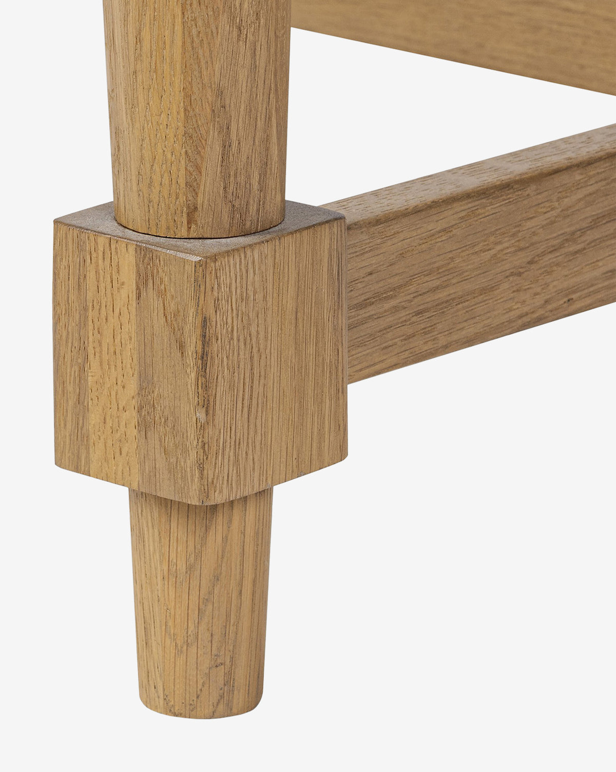 Close-up of a Kyler Oak Extension Dining Table leg with a square base and round, tapered lower section, joined to a horizontal support bar; all parts feature a light natural oak wood finish, by McGee & Co.
