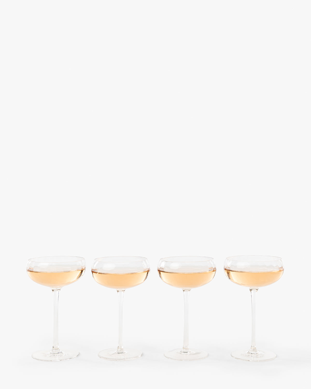 A set of four Langford Coupes, filled with a light pink sparkling beverage, are lined up against a plain white background, highlighting the elegance of this glassware set—perfect for any celebration, by McGee & Co.