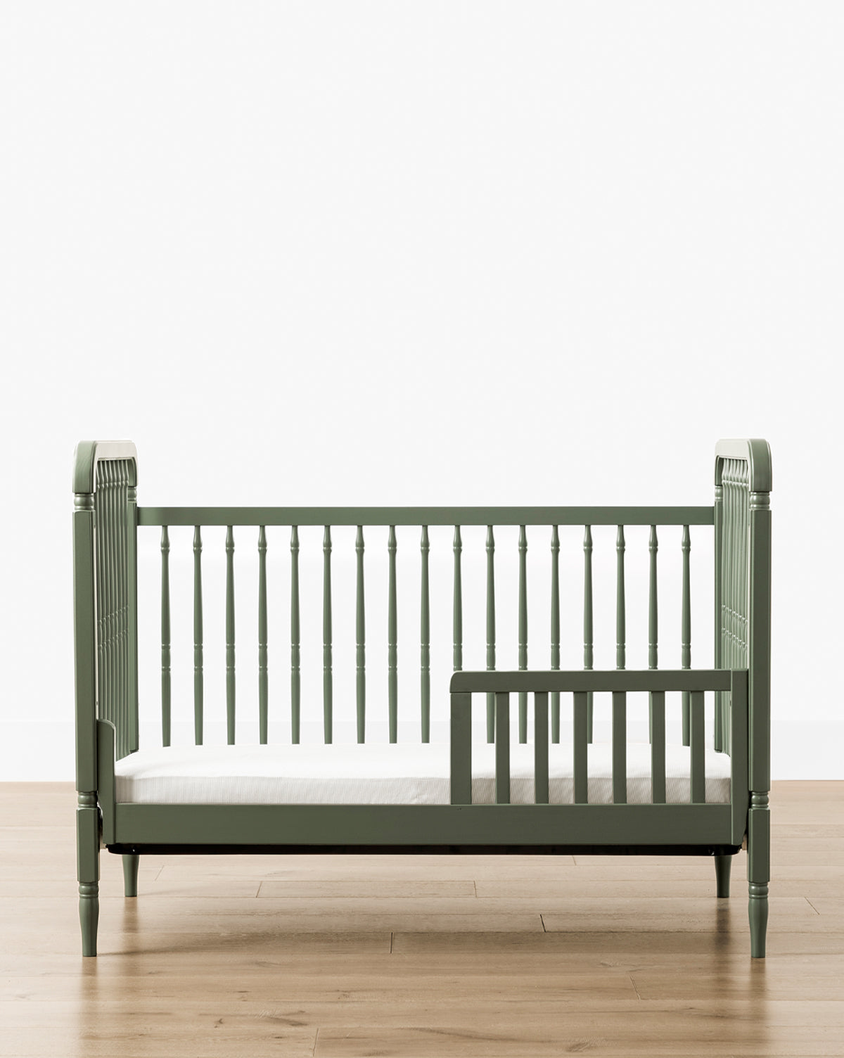 Liberty 3-in-1 Convertible Spindle Crib with Toddler Bed Conversion Kit in green, crafted from solid pine with vertical slats, low safety rail, and white mattress, shown on a light wood floor against a plain white background by McGee & Co.