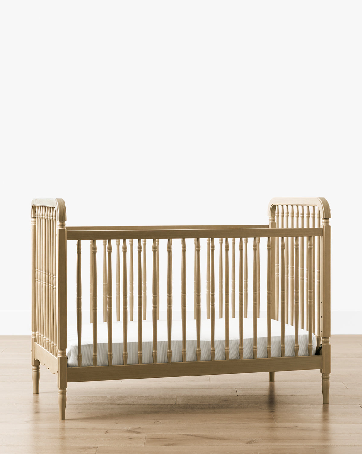 The Liberty 3-in-1 Convertible Spindle Crib w/Toddler Bed Conversion Kit, made of solid pine wood with vertical spindles and rounded corners, sits on a wooden floor against a plain white background and includes a thin mattress inside by McGee & Co.