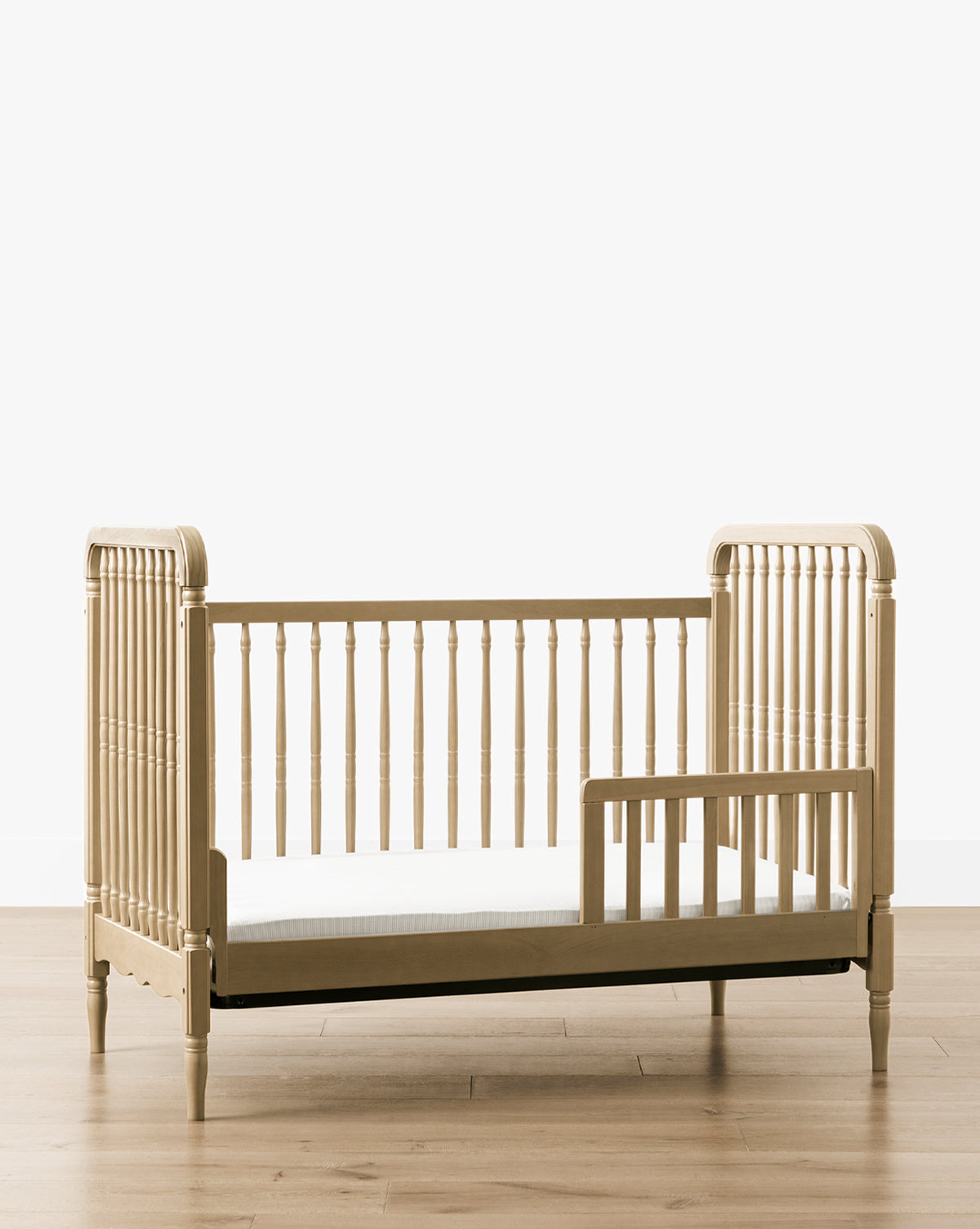 The Liberty 3-in-1 Convertible Spindle Crib w/Toddler Bed Conversion Kit, made from solid pine with vertical slats and a partial safety rail, is shown on a light wood floor against a plain white background by McGee & Co.