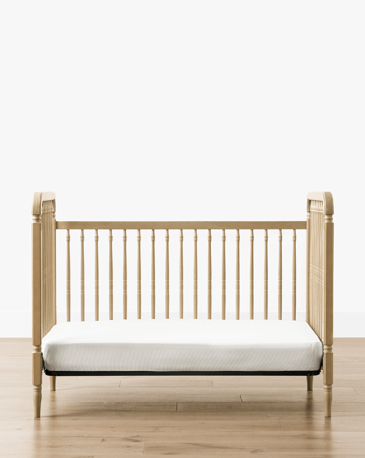 The Liberty 3-in-1 Convertible Spindle Crib w/Toddler Bed Conversion Kit, crafted from solid pine with vertical slats and rounded corners, features a white mattress and is set on a light wood floor against a plain white background, by McGee & Co.
