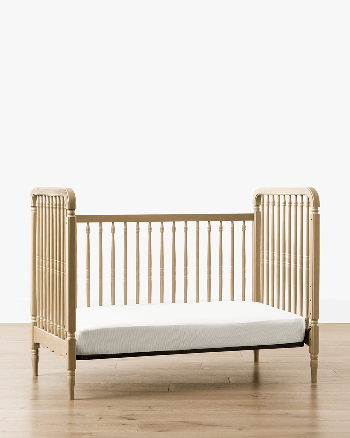 The Liberty 3-in-1 Convertible Spindle Crib w/Toddler Bed Conversion Kit becomes a light solid pine toddler bed with vertical side and back slats, plus a low, open front for easy access. It sits on a wooden floor against a plain white wall, by McGee & Co.