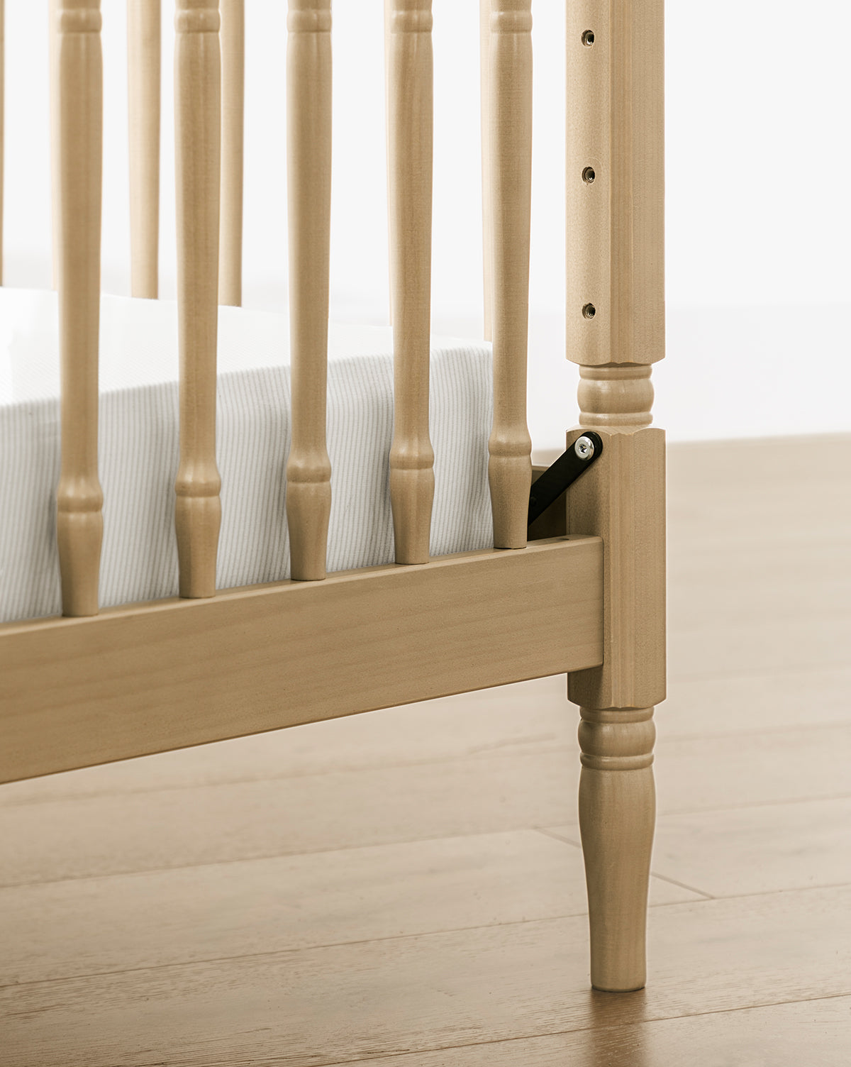 Close-up of the Liberty 3-in-1 Convertible Spindle Crib leg and side rail, made from solid pine, with vertical slats. A white mattress sits inside; height adjustment hardware is visible. The crib stands on a light wood floor, by McGee & Co.