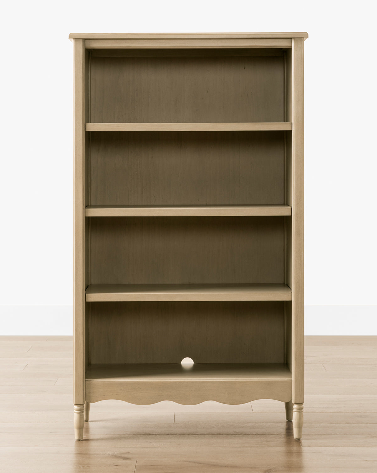 The Liberty Bookcase features three shelves and a scalloped bottom edge, set against a wooden floor. Its dark back panel with a small round hole adds distinctive character to this timeless piece, by McGee & Co.
