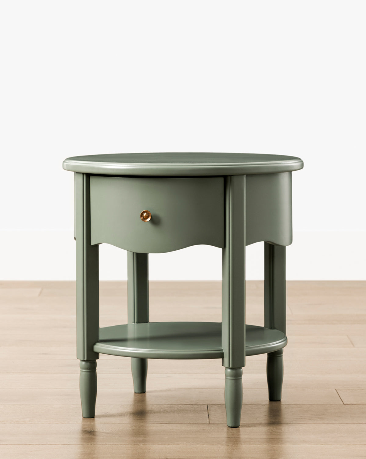 The Liberty Nightstand is a round, green piece with a single drawer and brass knob, lower shelf, and four turned legs. It sits on a light wooden floor against a white wall. GREENGUARD Gold Certified for safer indoor air quality, by McGee & Co.
