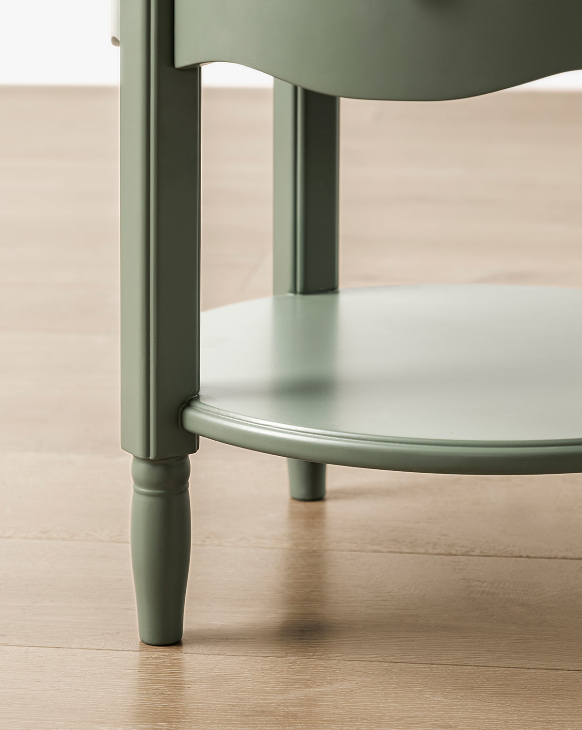 Close-up of the lower section of the GREENGUARD Gold Certified Liberty Nightstand, featuring a green round top, slender legs, and a circular lower shelf—a safe and stylish addition to any nursery on a light wood floor, by McGee & Co.