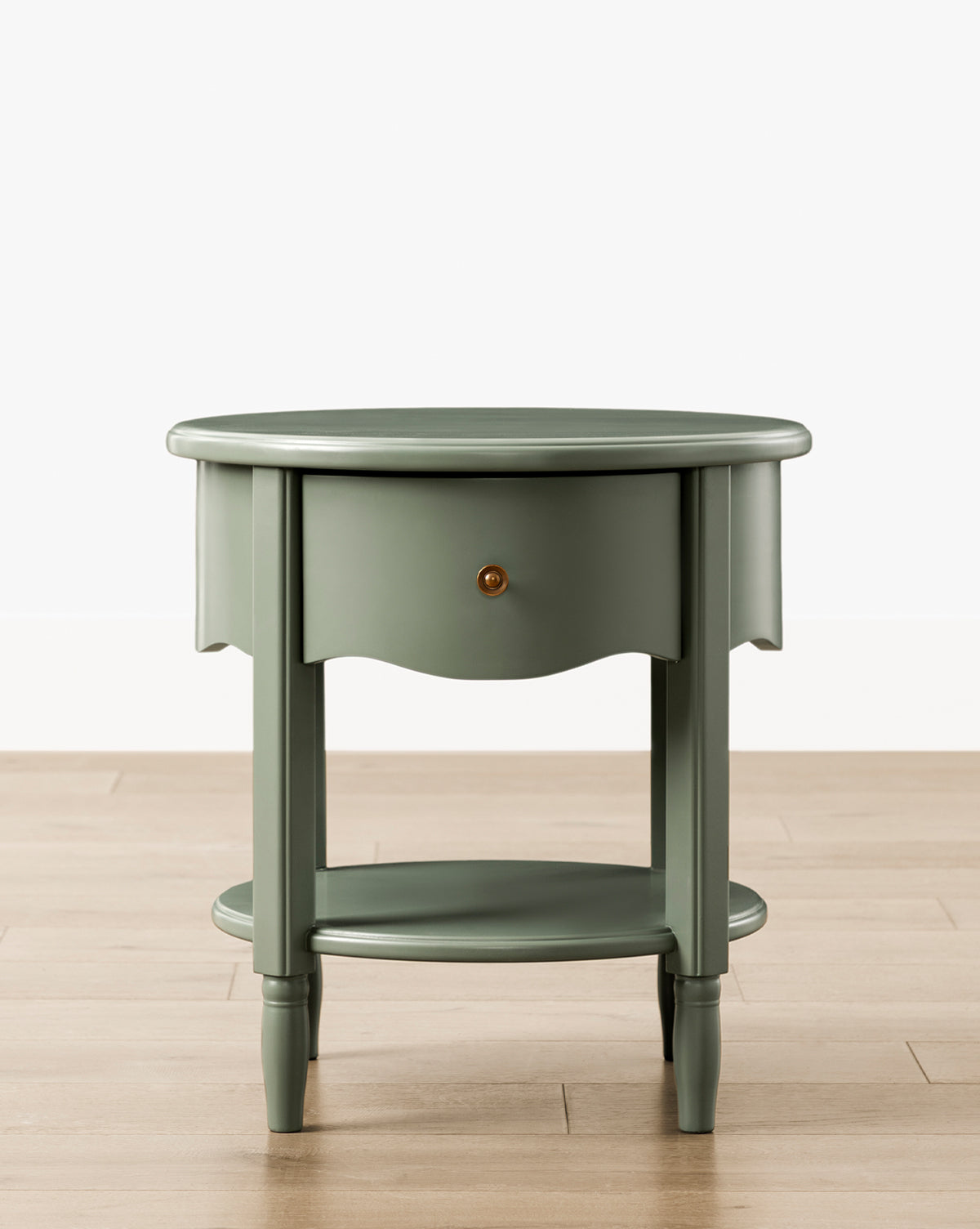 The Liberty Nightstand, a round, sage green piece with a small drawer, lower shelf, and four tapered legs, sits on a light wood floor against a white background. GREENGUARD Gold Certified—perfect for nurseries and healthier indoor air by McGee & Co.