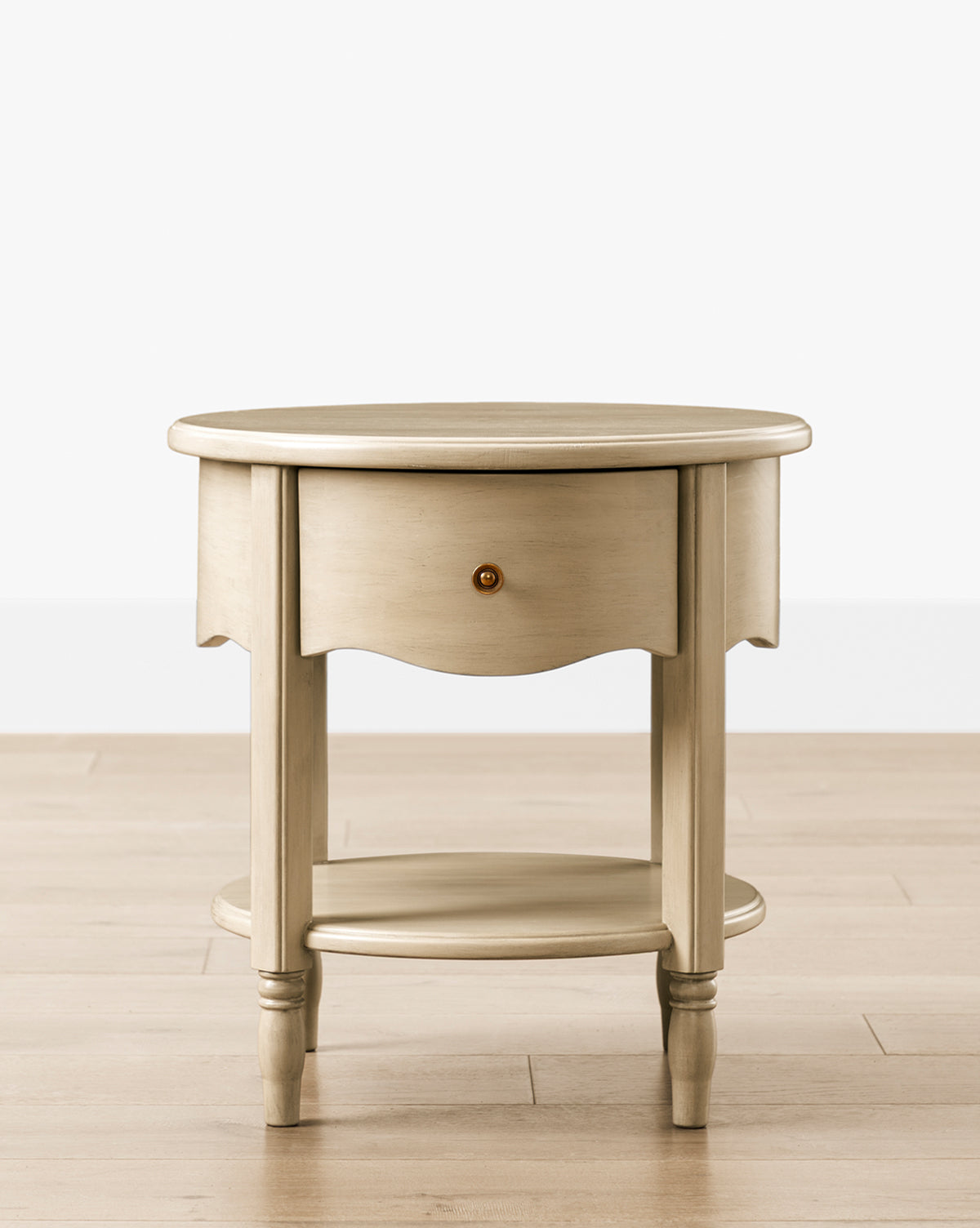 The Liberty Nightstand is a round wooden table with a light, distressed finish, one drawer with a round knob, four legs, and a lower shelf. It sits on a light wood floor against a white background and is GREENGUARD Gold Certified for nurseries by McGee & Co.