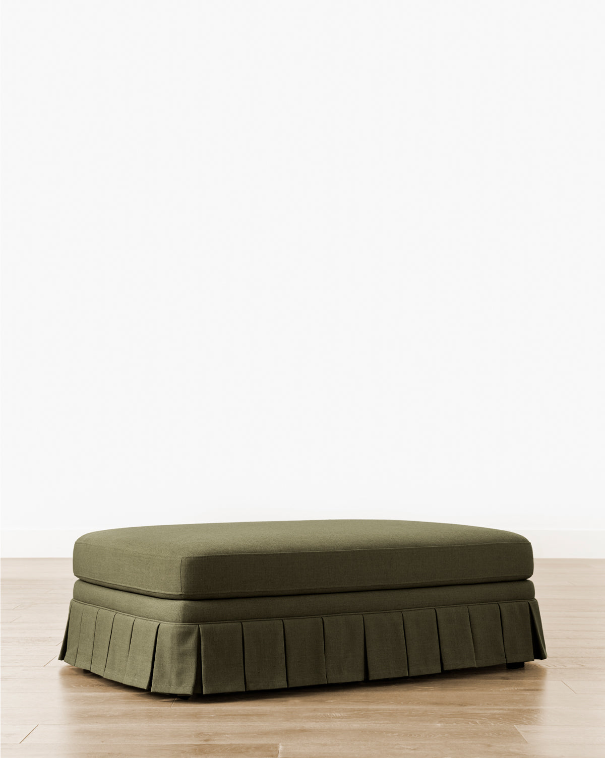 The Louisa Ottoman showcases custom olive green upholstery and a box pleat skirt, displayed elegantly on a light wood floor with a plain white background, by McGee & Co.
