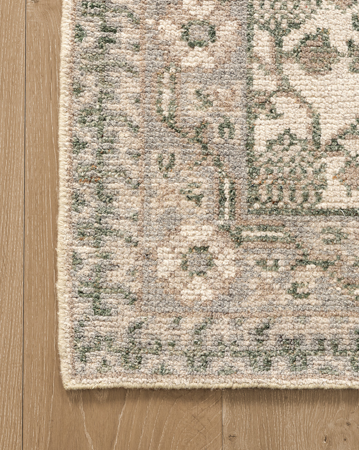 Close-up of the corner of the Orsino Hand-Knotted Rug in green, beige, and cream on a light wood floor, showcasing its antique style with a decorative border featuring floral and geometric motifs, by McGee & Co.