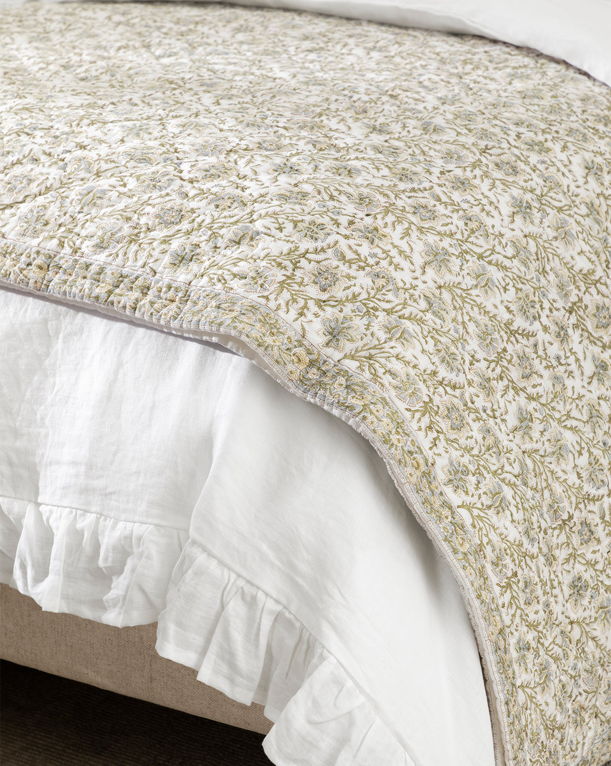 A close-up of a bed featuring the Seton Kids Quilt, showcasing its delicate green and beige floral motif and a white ruffled bed skirt draped gracefully over the edge, by McGee & Co.