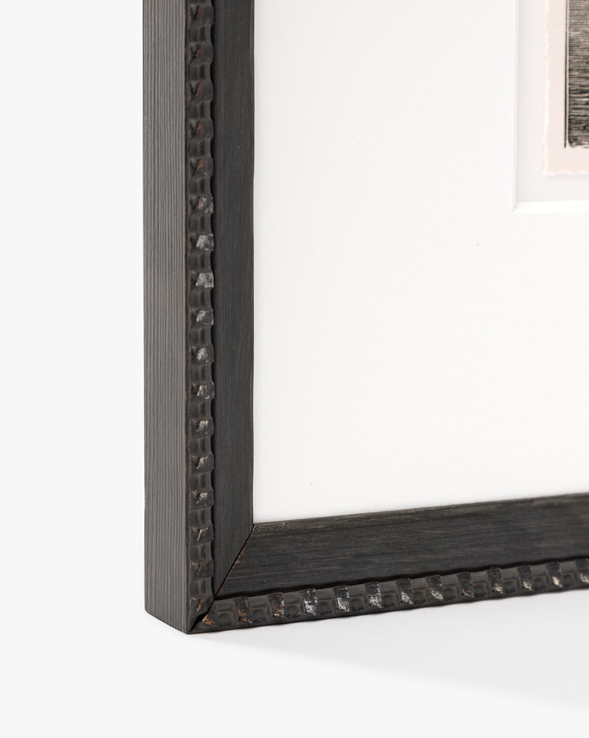 Close-up of the bottom left corner of a black, hammered metal-look frame with minimalist design and white mat board. The upper right reveals part of a seascape sketch, inspired by Shoals Engraving, by McGee & Co.