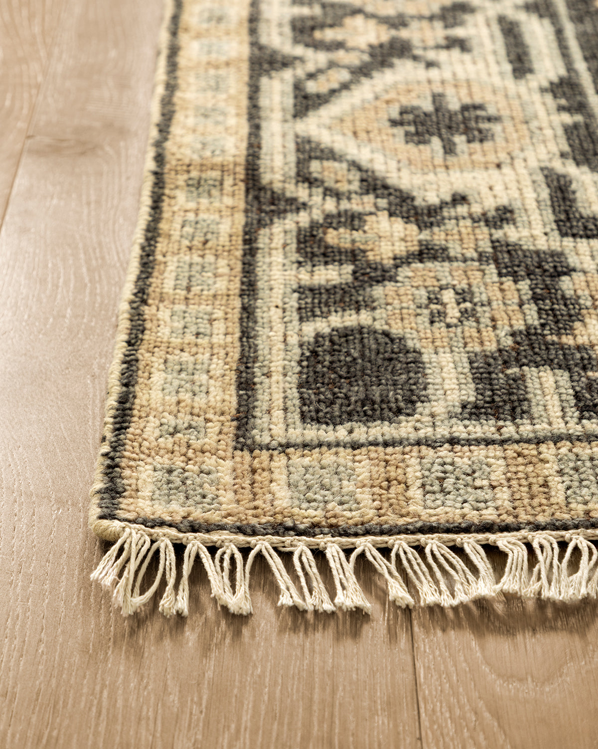 A close-up of the Wayland Hand-Knotted Wool Rug, showing beige, black, and cream patterns with fringe along the edge, laid on a light wooden floor, by McGee & Co.