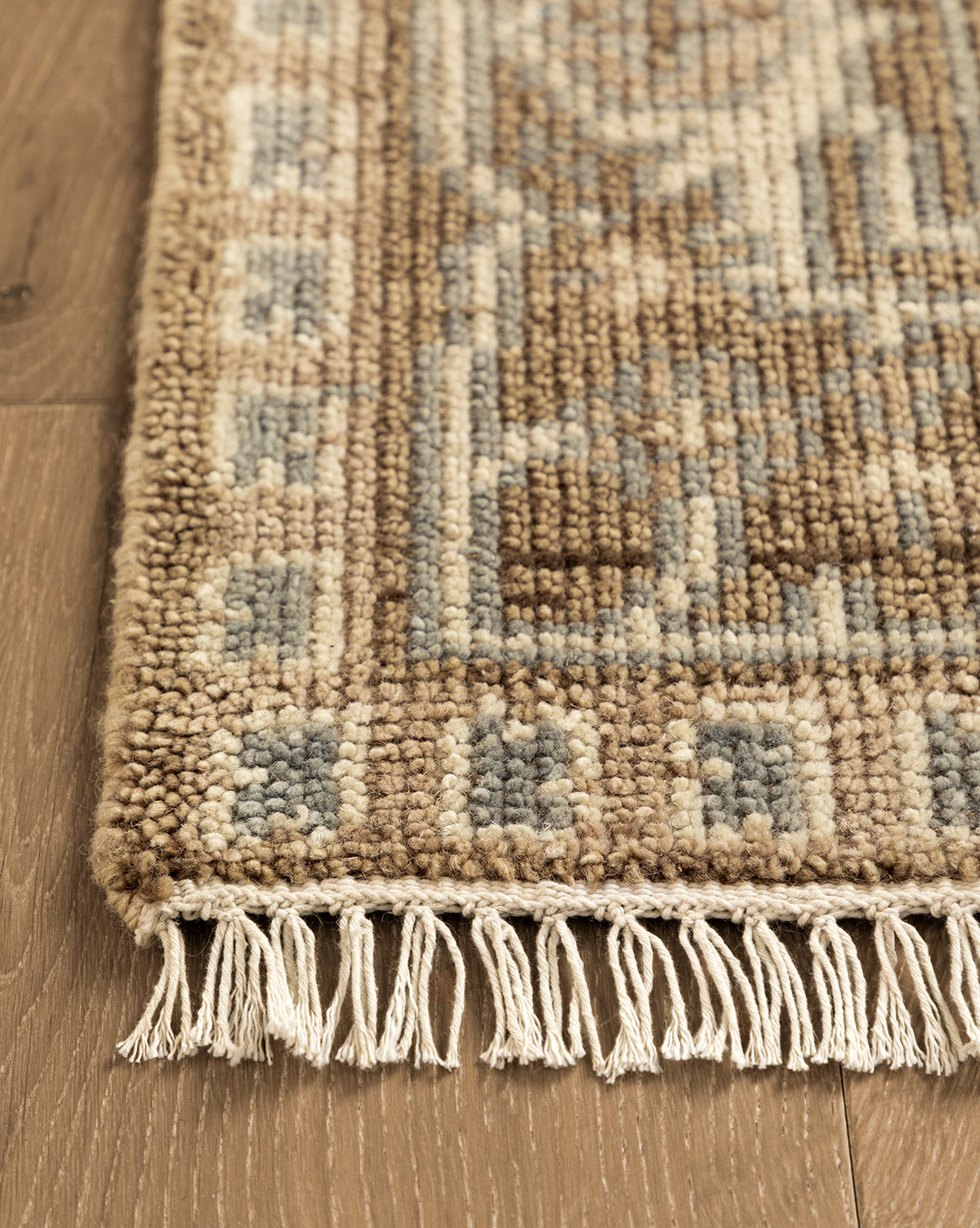 A close-up of the corner of the Wayland Hand-Knotted Wool Rug in beige, brown, and blue with knotted fringe on a wooden floor—highlighting its artisan craftsmanship, by McGee & Co.