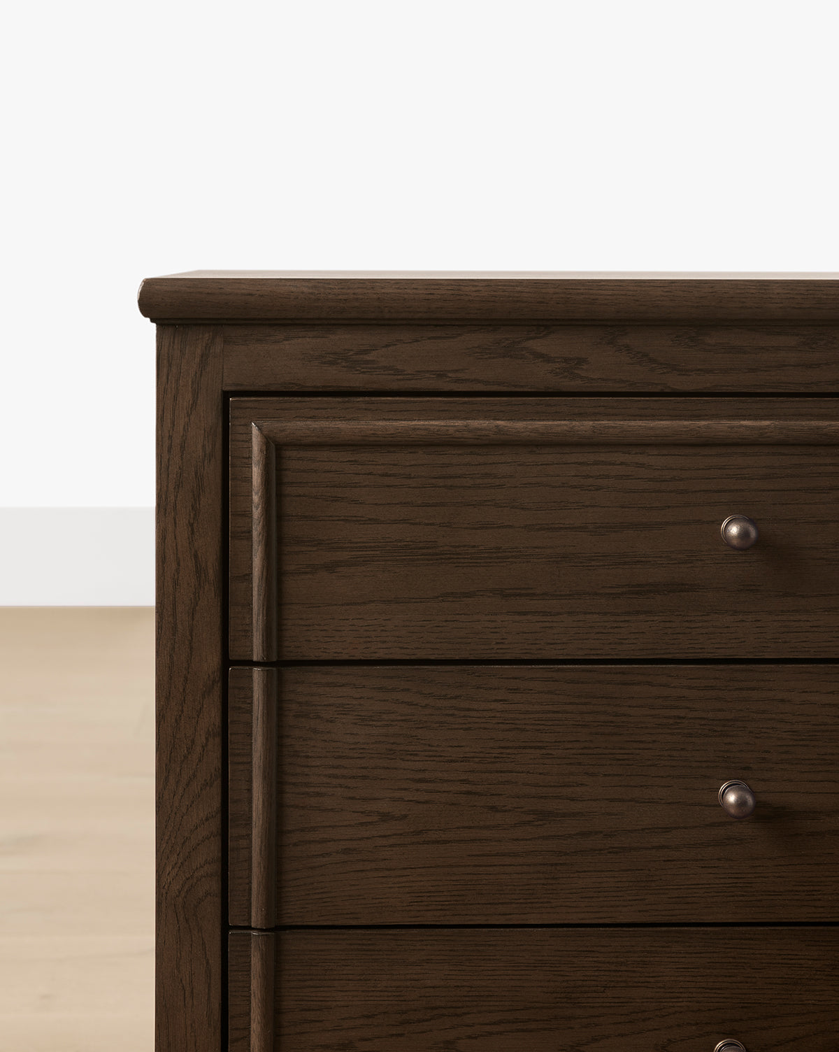 A close-up of the Yorkshire Nightstand’s top left corner reveals its English oak finish, three drawers with round antique brass pulls, and a dark wooden build set against a light floor and white wall, by McGee & Co.