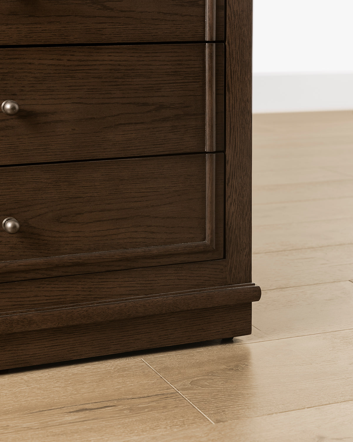 Close-up of the bottom corner of the Yorkshire Nightstand in an English oak finish with antique brass pulls, placed on a light wood floor against a white wall by McGee & Co.