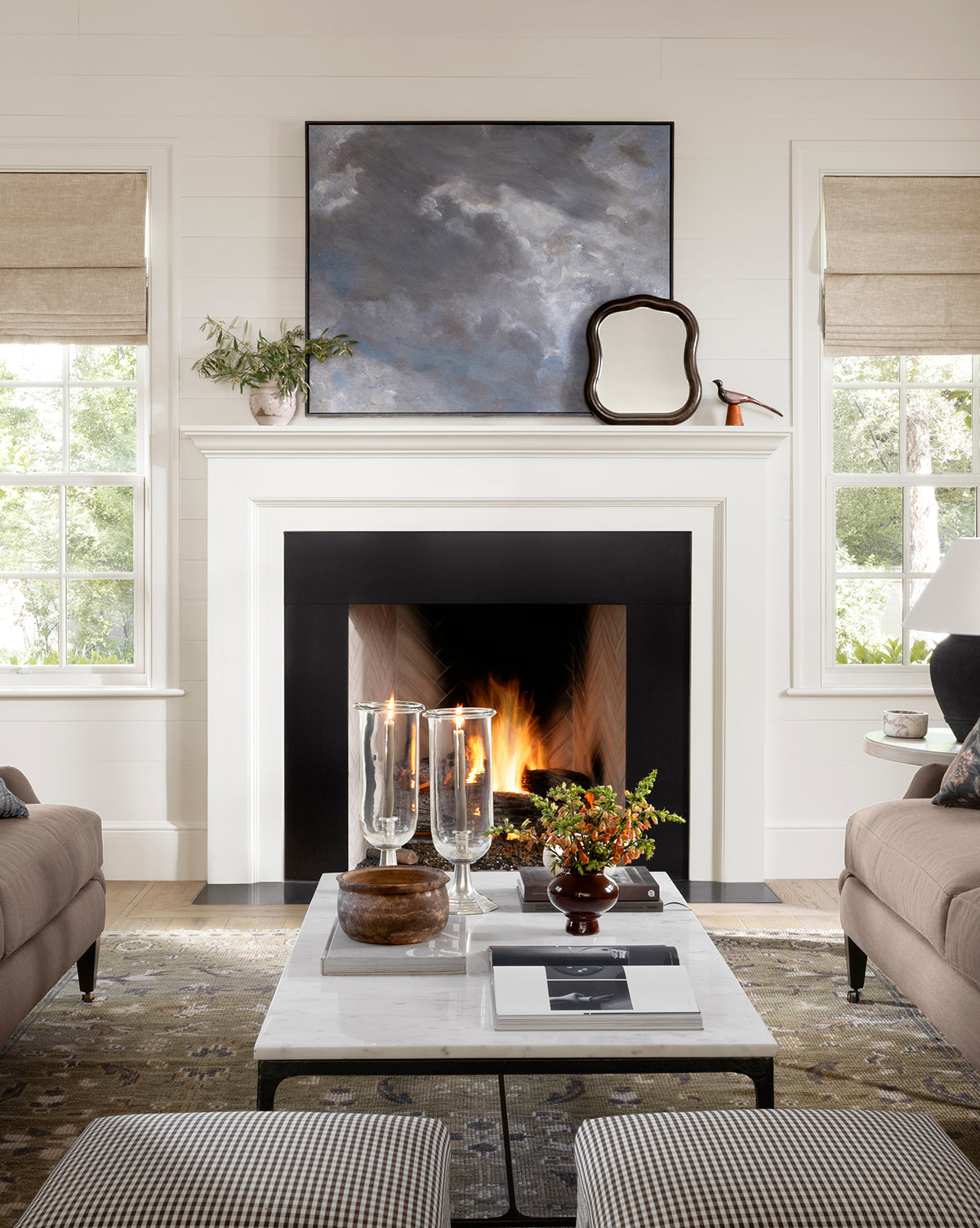 A cozy living room with a lit fireplace, Dark Skies Ahead giclée canvas in a floater frame and mirror above the mantel, two windows, beige sofas, and a white marble coffee table decorated with candles, books, and a small centerpiece by McGee & Co.