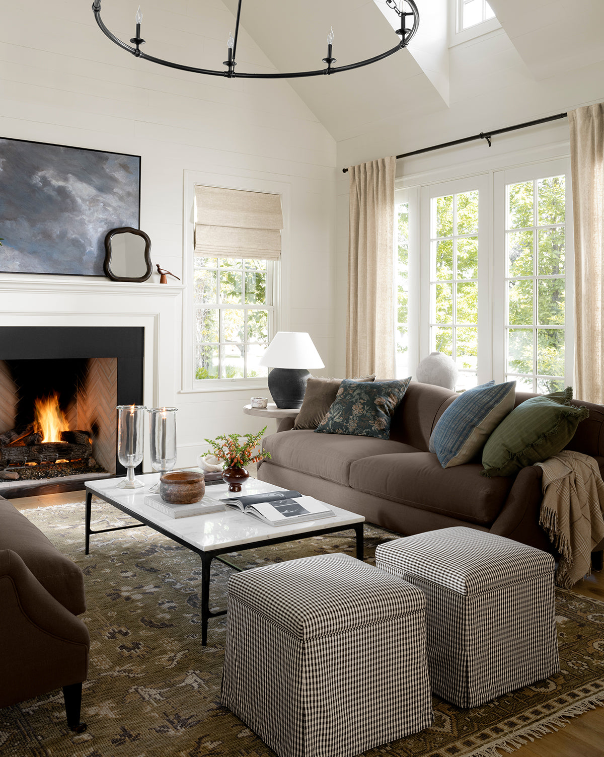 A cozy living room with a lit fireplace, brown sofas accented by green and blue pillows, a white marble coffee table, two Latimer Ottomans (Ready to Ship), a large rug, and beige curtains framing sunlit windows—ideal for home styling enthusiasts, by McGee & Co.