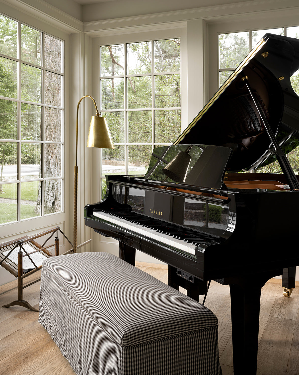 A black Yamaha grand piano sits in a bright room with large windows, a Latimer Bench in front, a brass floor lamp beside it, and trees visible outside, by McGee & Co.