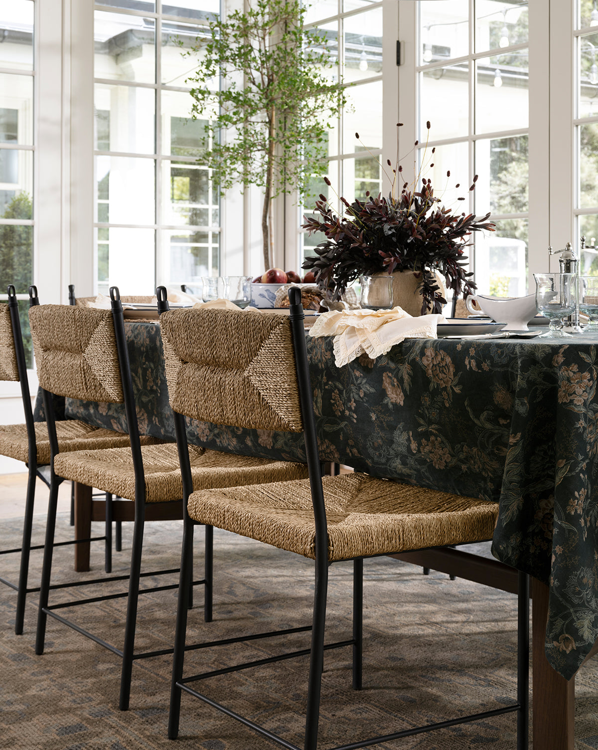 A dining table set with a dark floral tablecloth, Peregrine Chairs, glassware, plates, and a centerpiece of dark foliage in a bright room with large windows and a potted tree by McGee & Co.