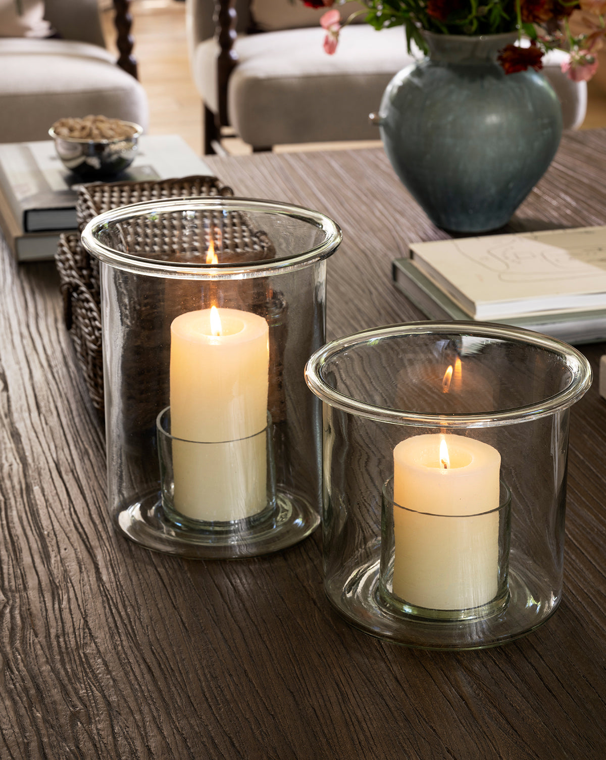 Two lit pillar candles in Cyrus Hurricane holders sit on a wooden table. Nearby are a ceramic vase with flowers, stacked books, a woven basket, and snacks, creating a cozy, inviting home decor atmosphere by McGee & Co.