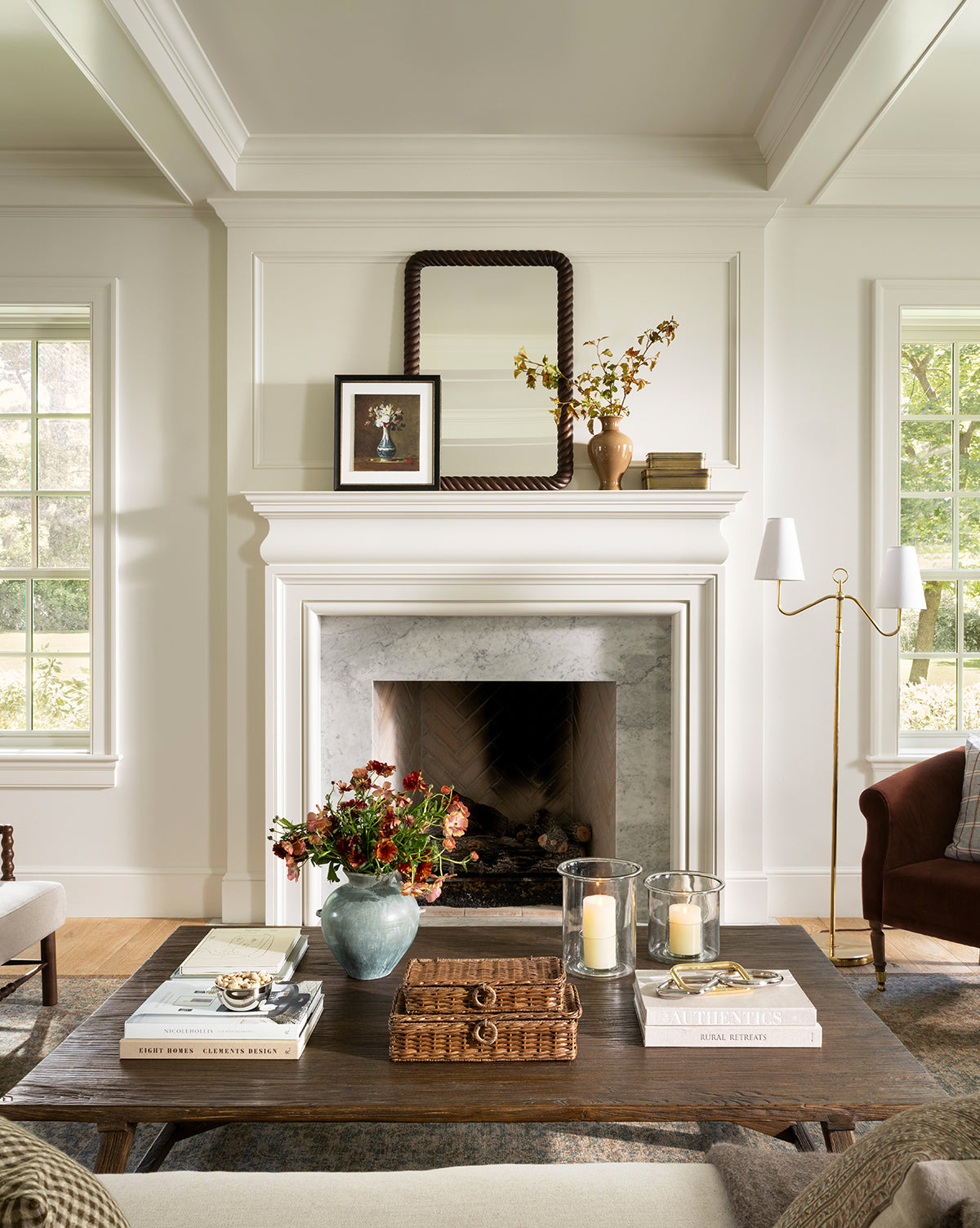 Elegant living room with a white fireplace, Mayweather Wall Mirror with a rope-inspired frame above the mantel, framed art, and a vase with branches. A dark red walnut coffee table displays books, candles, flowers, and a wicker tray. Sunlight streams through large windows, by McGee & Co.
