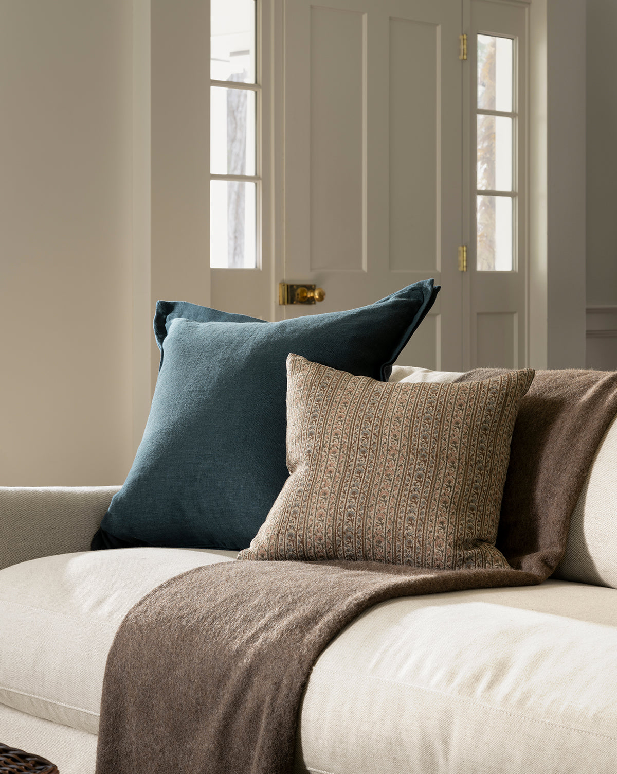 A beige sofa with a dark blue pillow, a patterned brown pillow, and the Locester Throw sits in a bright room with light walls and a glass-paneled door in the background, by McGee & Co.