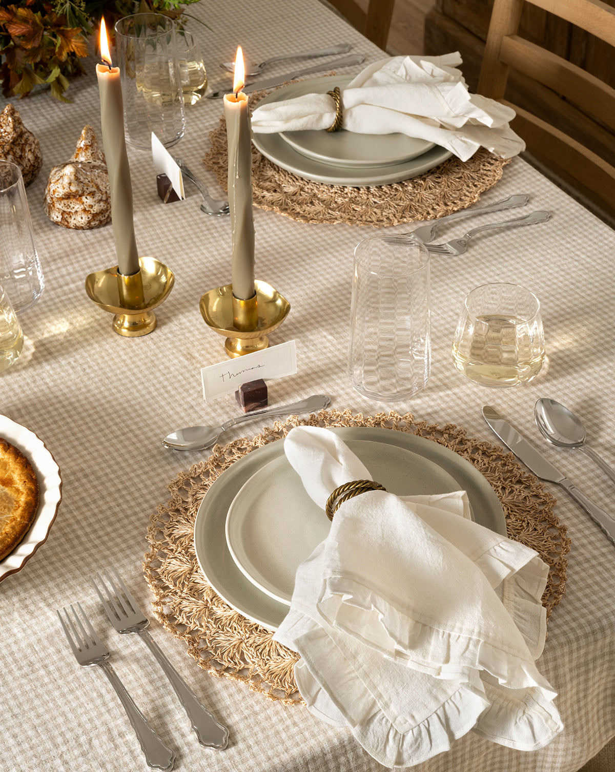 A set dining table with neutral-toned plates, white cloth napkins and rings, woven placemats, clear glasses, the Artois Flatware (Set of 5), brass candlesticks with lit candles, place cards, and a checked tablecloth by McGee & Co.
