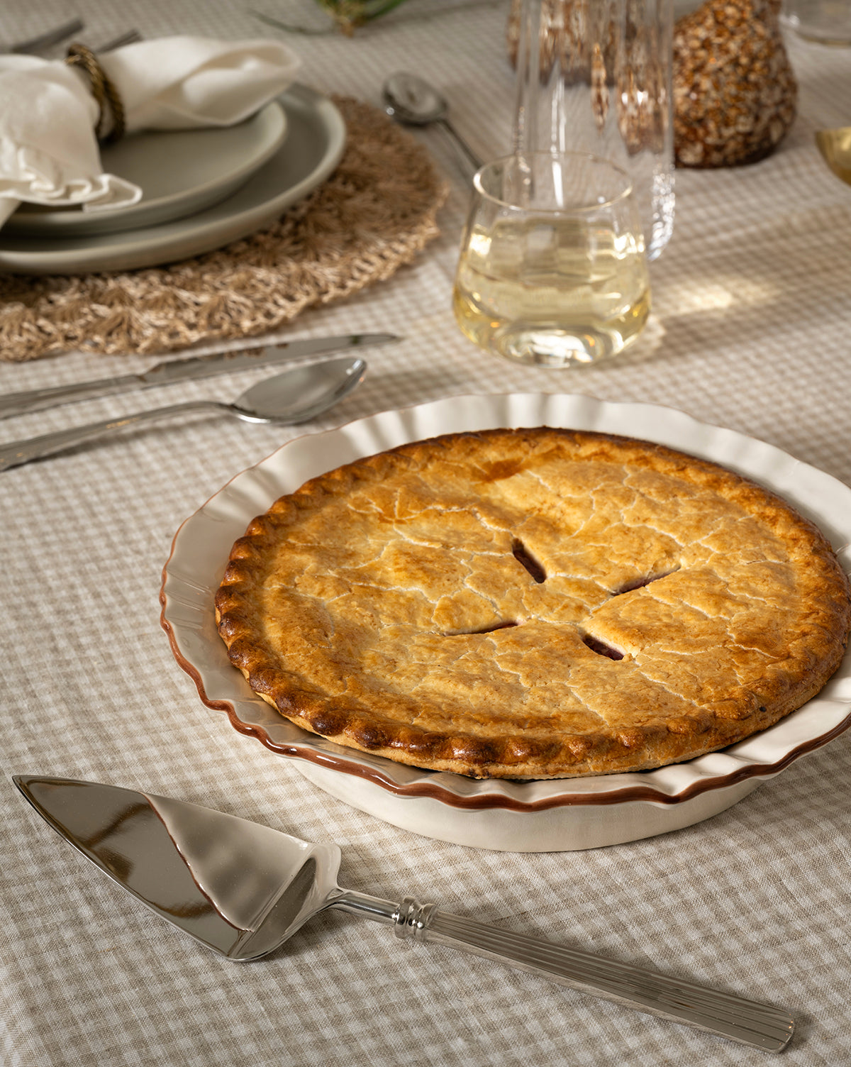 A golden-brown pie rests in a white ceramic dish on a checked tablecloth, accompanied by the McNully Pie Server, silver cutlery, a glass of white wine, and a neatly set plate with a folded napkin by McGee & Co.