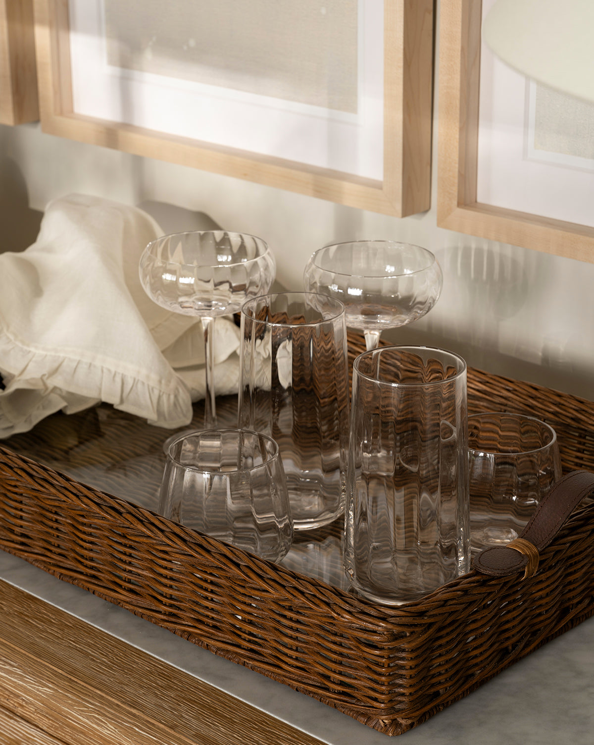A brown wicker tray holds several Langford Highball Glasses (Set of 4) and folded cloth napkins on a marble surface, with framed pictures in the background, by McGee & Co.